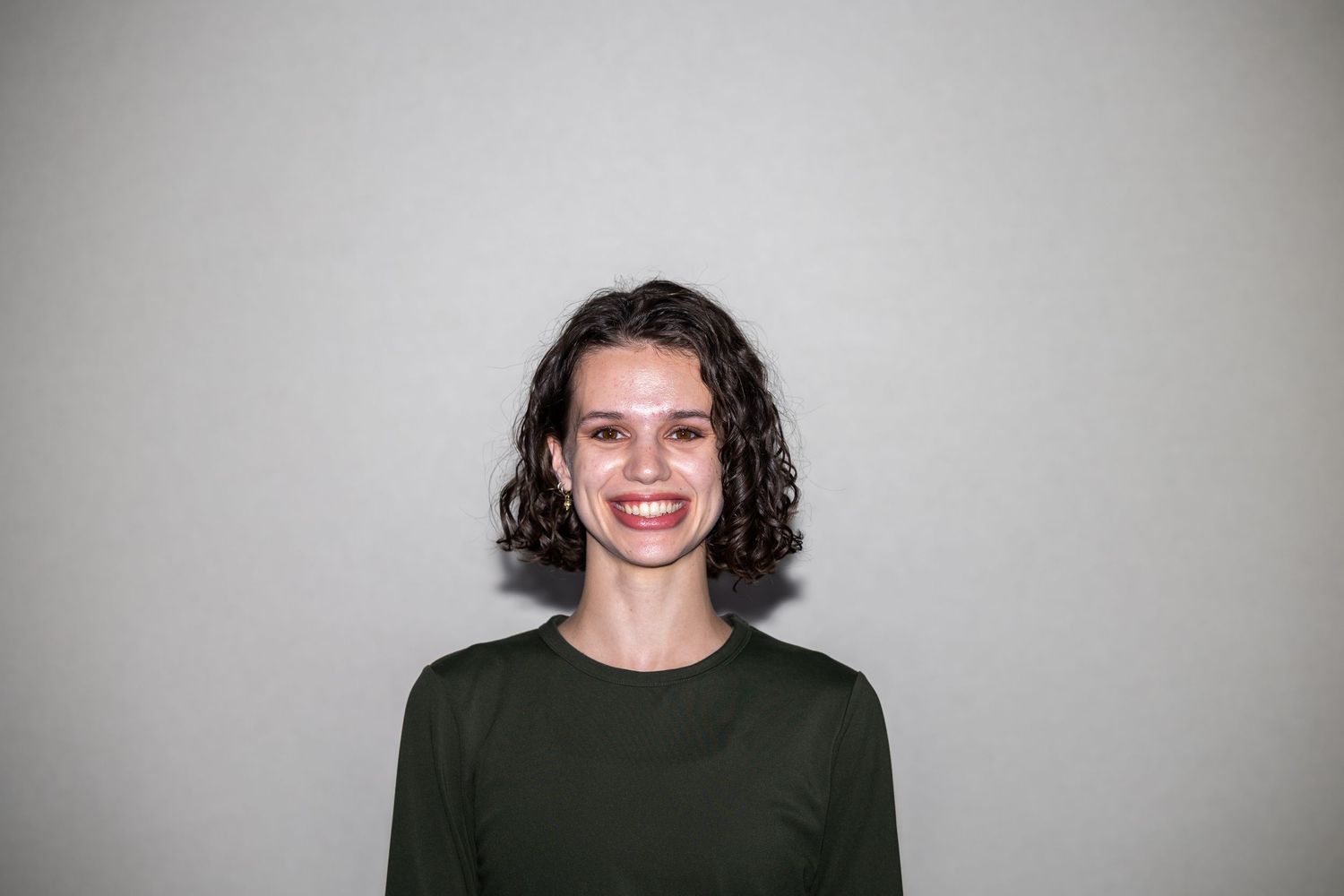 Smiling young woman with short curly dark hair wearing a dark green long-sleeve top against a gray background.