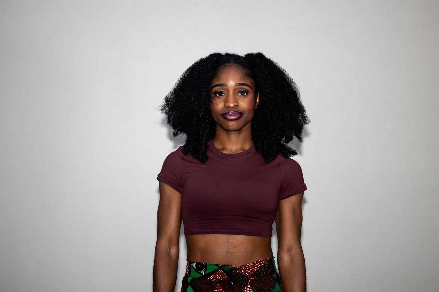 Young Black woman with curly hair wearing a maroon crop top and colorful printed skirt posing against white background.