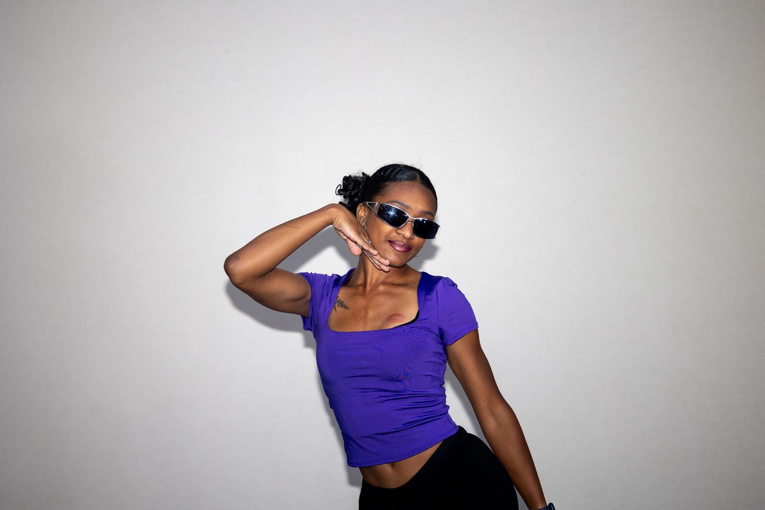 Stylish young Black woman in a purple crop top wearing sunglasses posing confidently against a gray background.