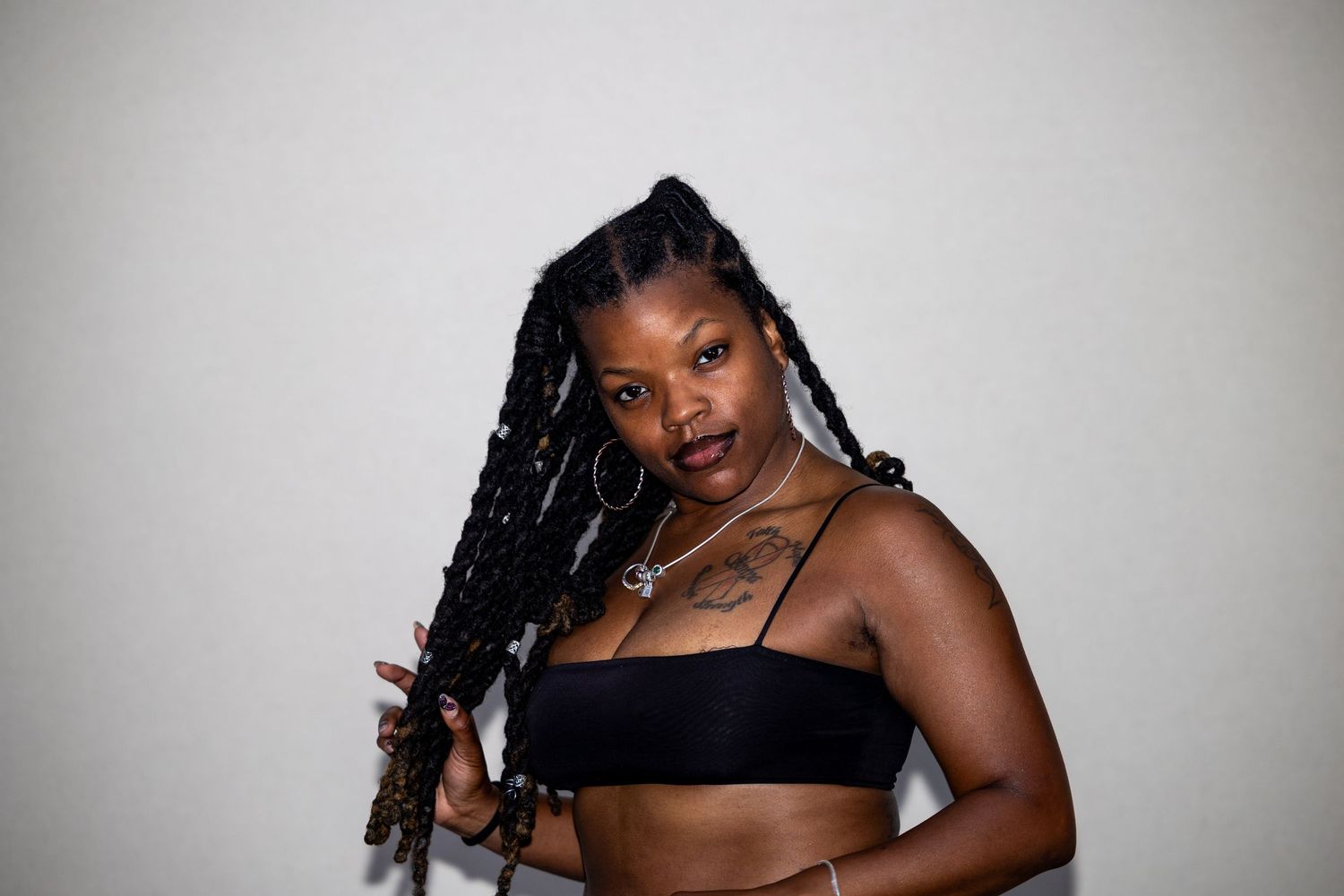 Young woman with long braids and hoop earrings in black crop top poses against white background.