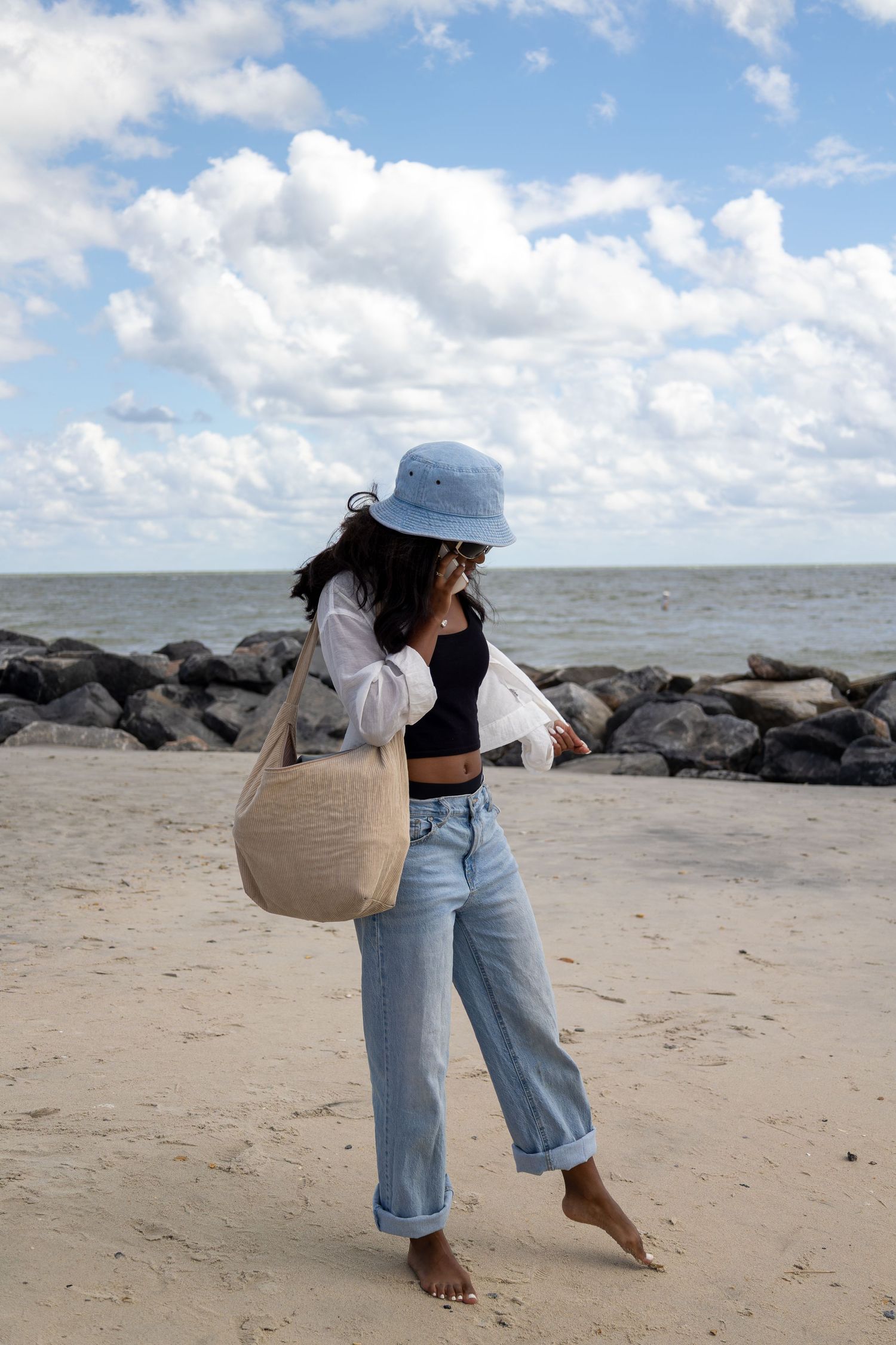 Someone in casual denim and a sun hat walks along a rocky beach shoreline.