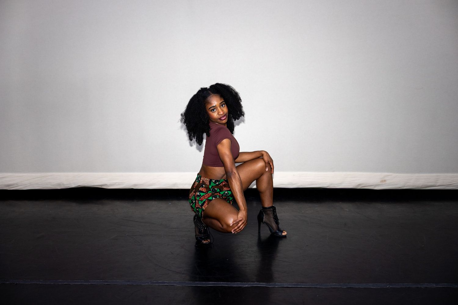 Woman in burgundy top and printed skirt kneeling gracefully on black studio floor.