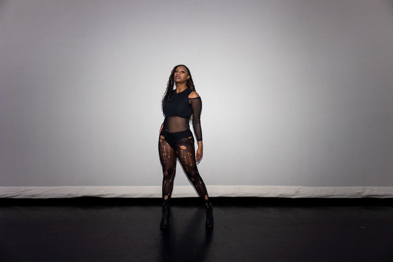 Woman in black sheer outfit posing confidently against a gray studio background.