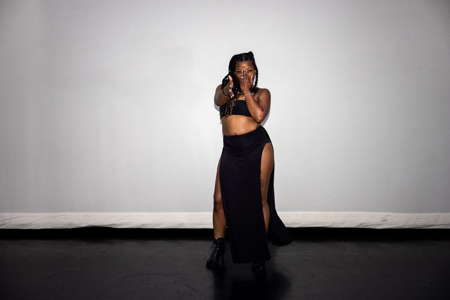 A woman in a black crop top and long split skirt poses dramatically covering her face against a gray background.