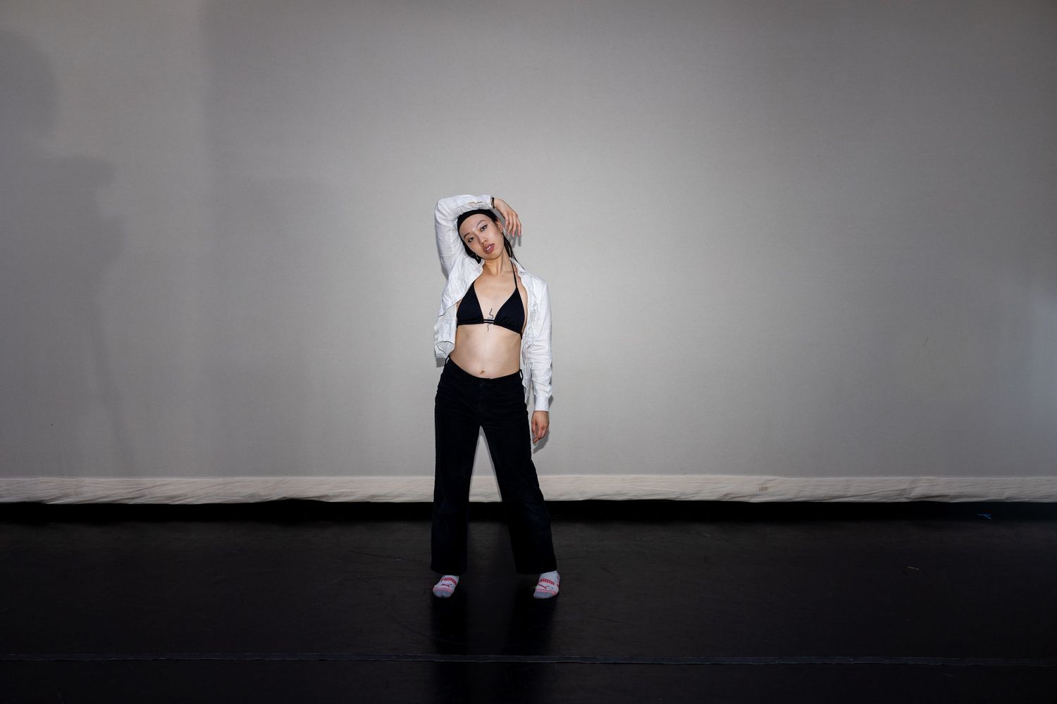 A woman in a black sports bra, black pants, and white cardigan stretches gracefully in a minimalist studio setting.