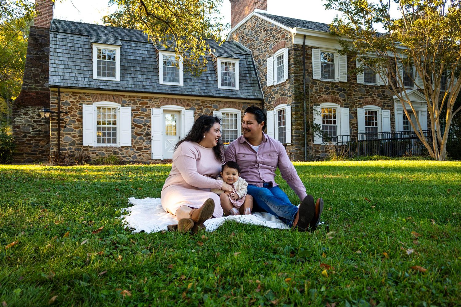 Family of three sitting on white blanket on grass in front of historic stone manor house surrounded by trees.