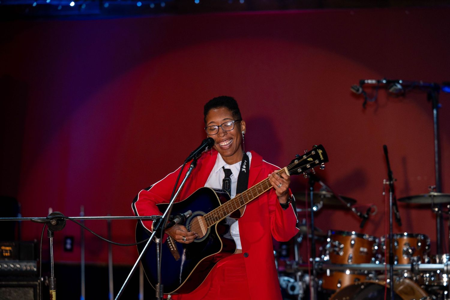 Smiling musician in red jacket playing acoustic guitar on stage at a live concert performance.