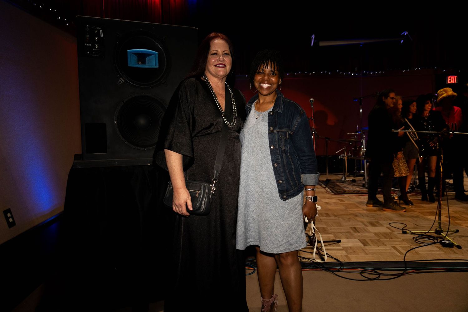 Two women smiling and posing together at an indoor music venue event with a band performing in the background.