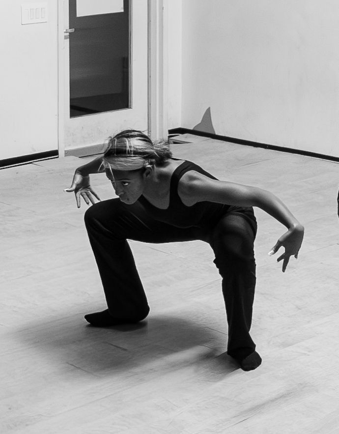 Black and white photo of a dancer in a low crouch pose, wearing all black, performing in a studio.