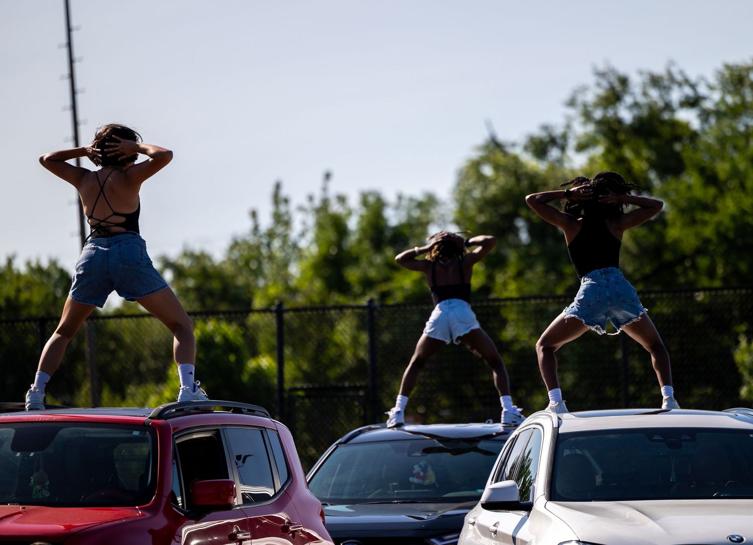 Three people dancing on top of cars in a parking lot with trees in the background on a sunny day.