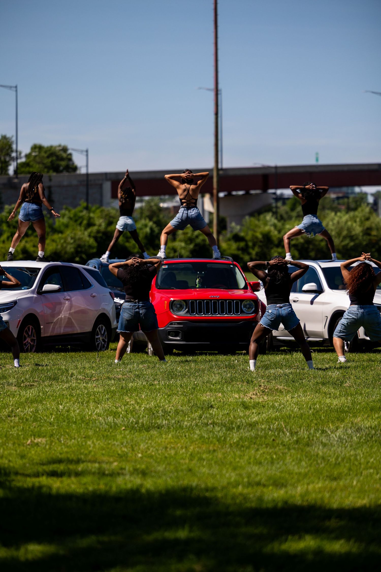 Shirtless male dancers performing synchronized routine on cars in a parking lot on a sunny day.
