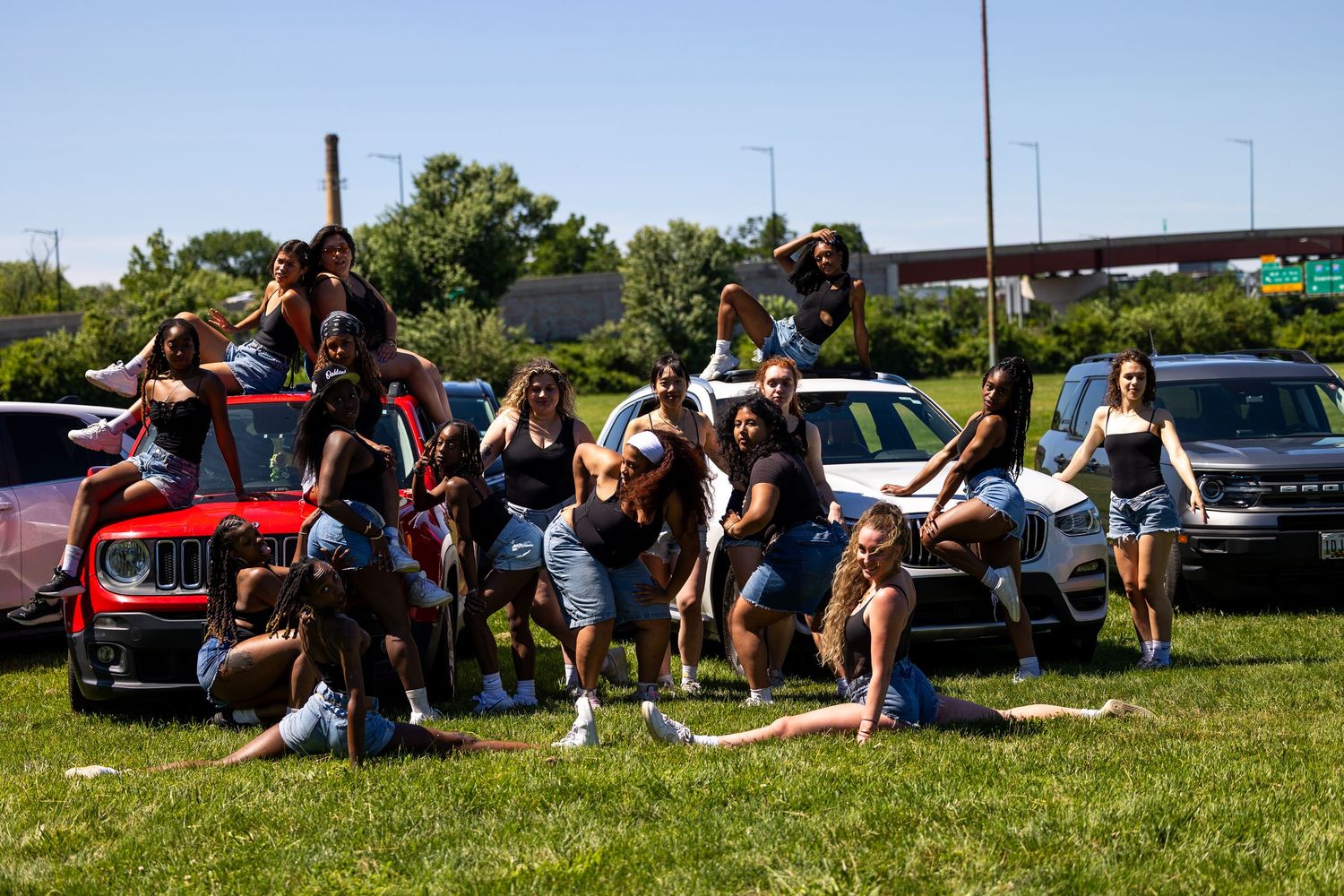 Group of young women posing together in a parking lot near cars on a sunny day.