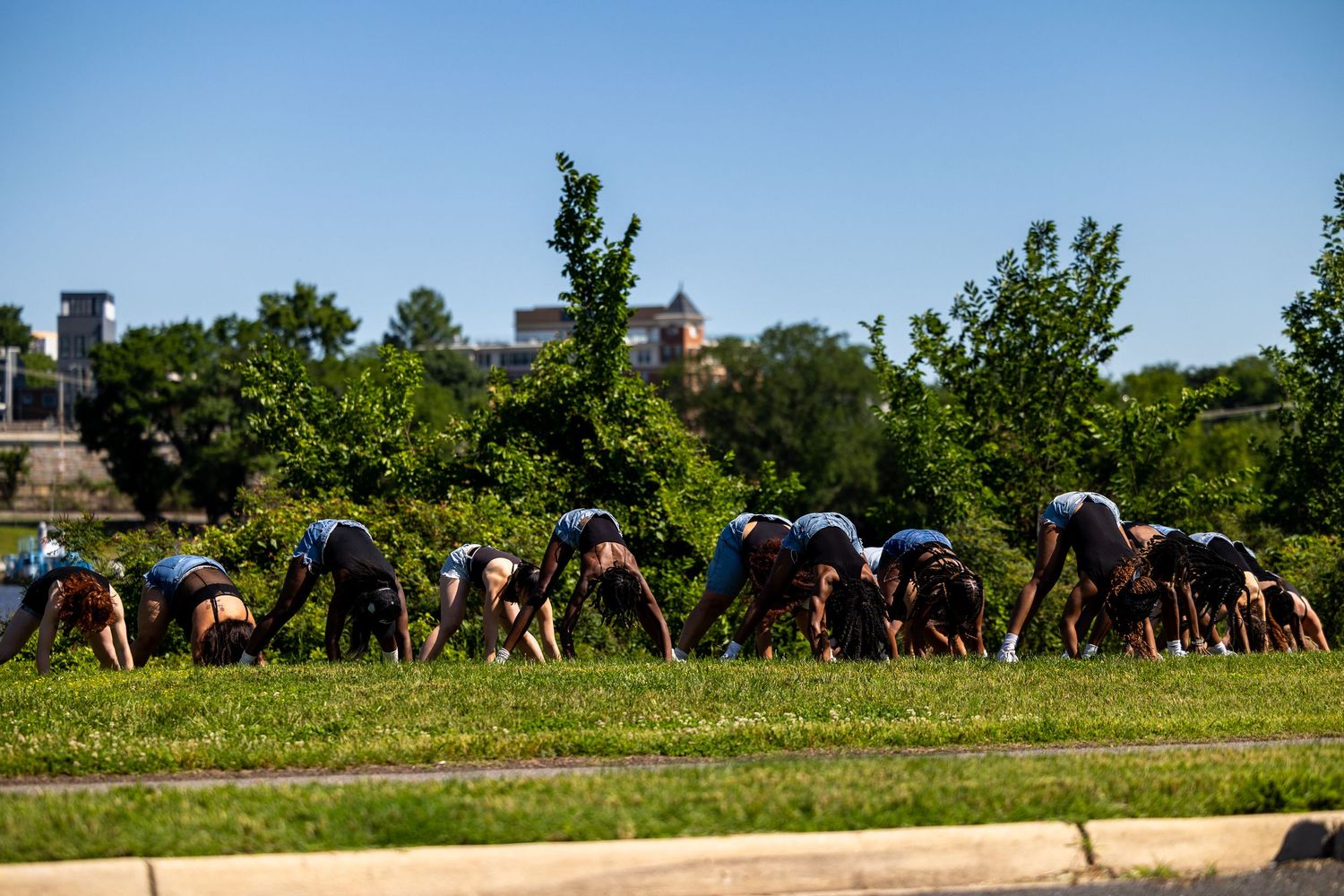 Group of people doing downward dog yoga pose outdoors on green grass with trees and building in background.