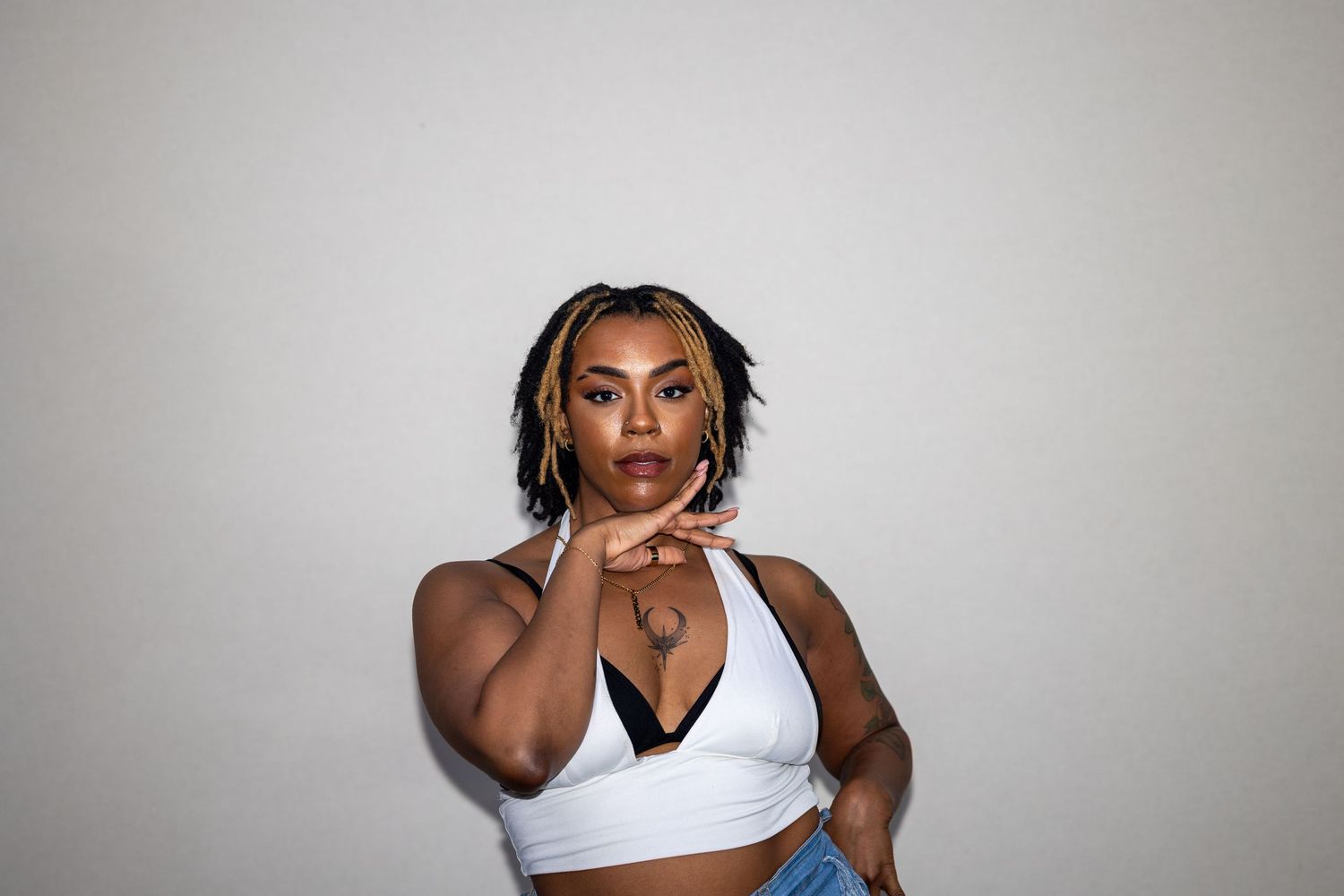 Confident woman in white crop top and jeans posing with hands clasped against a light gray background.