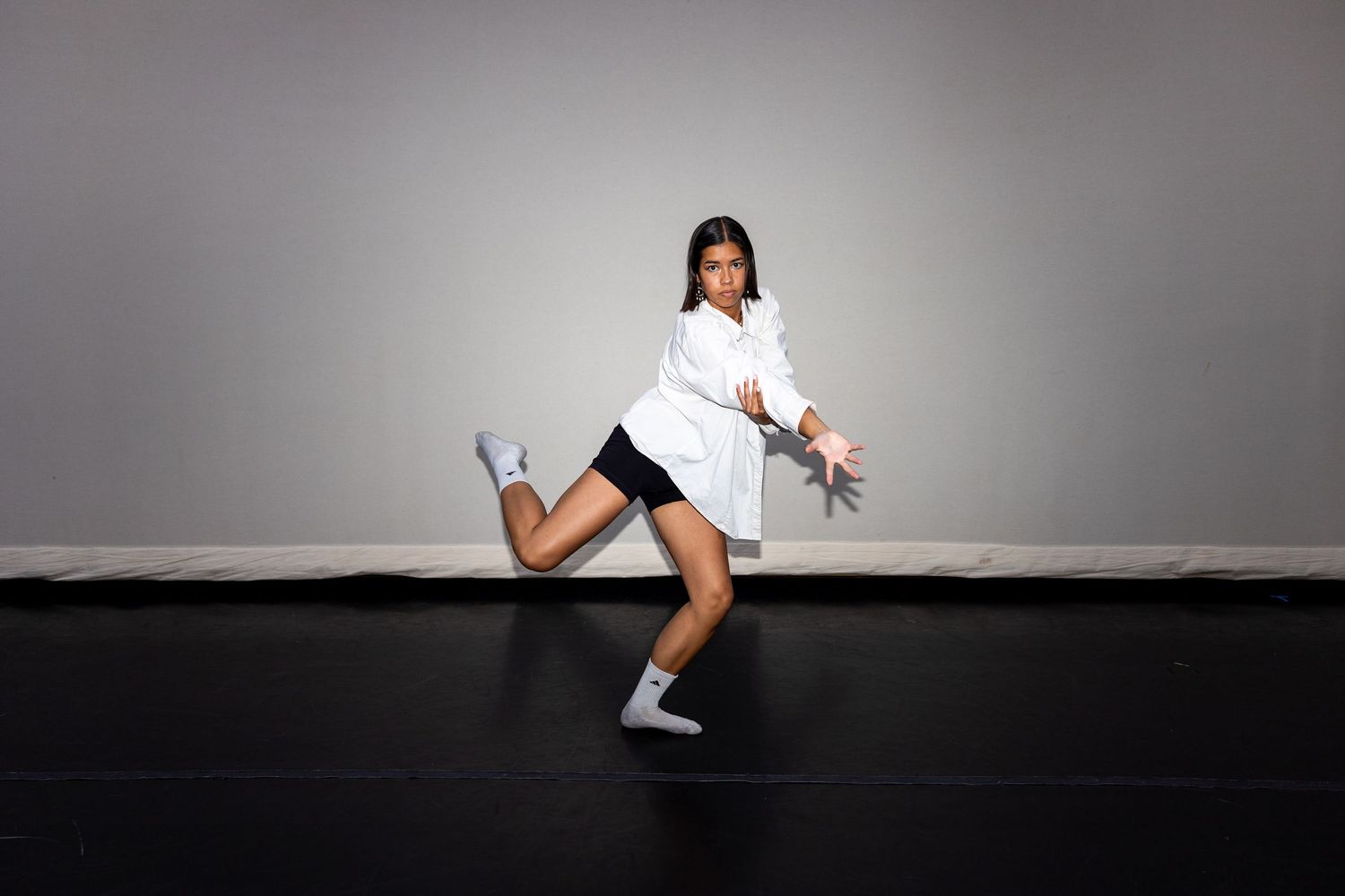 A dancer in white shirt and black shorts strikes a dynamic pose on a black floor against a gray background.