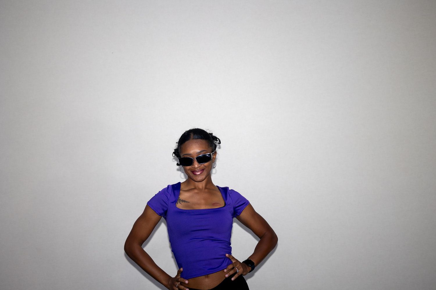 A stylish woman in a purple square-neck top and sunglasses poses with hands on hips against a white background.