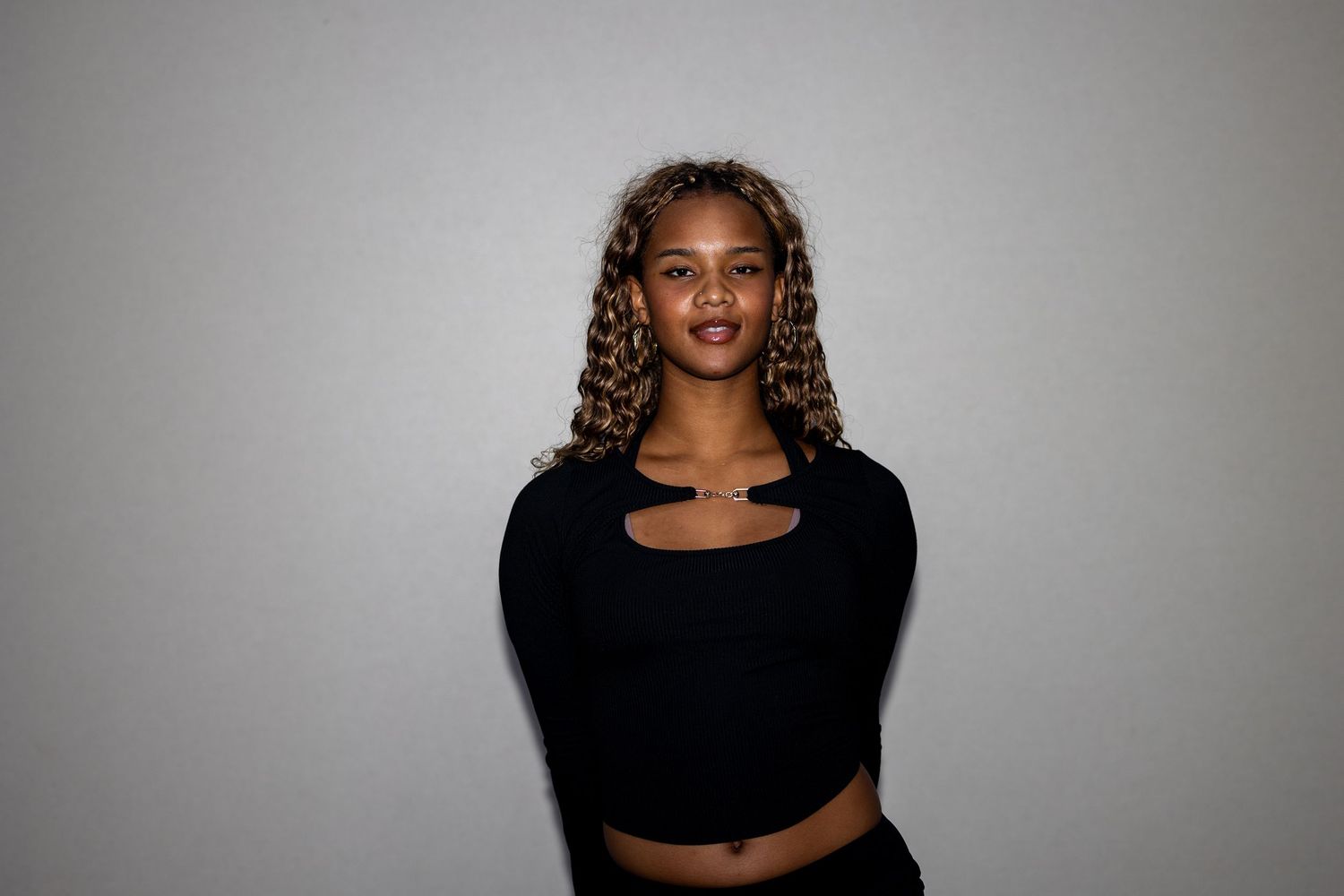 Woman with blonde curly hair wearing a black long-sleeve crop top and black pants poses calmly.