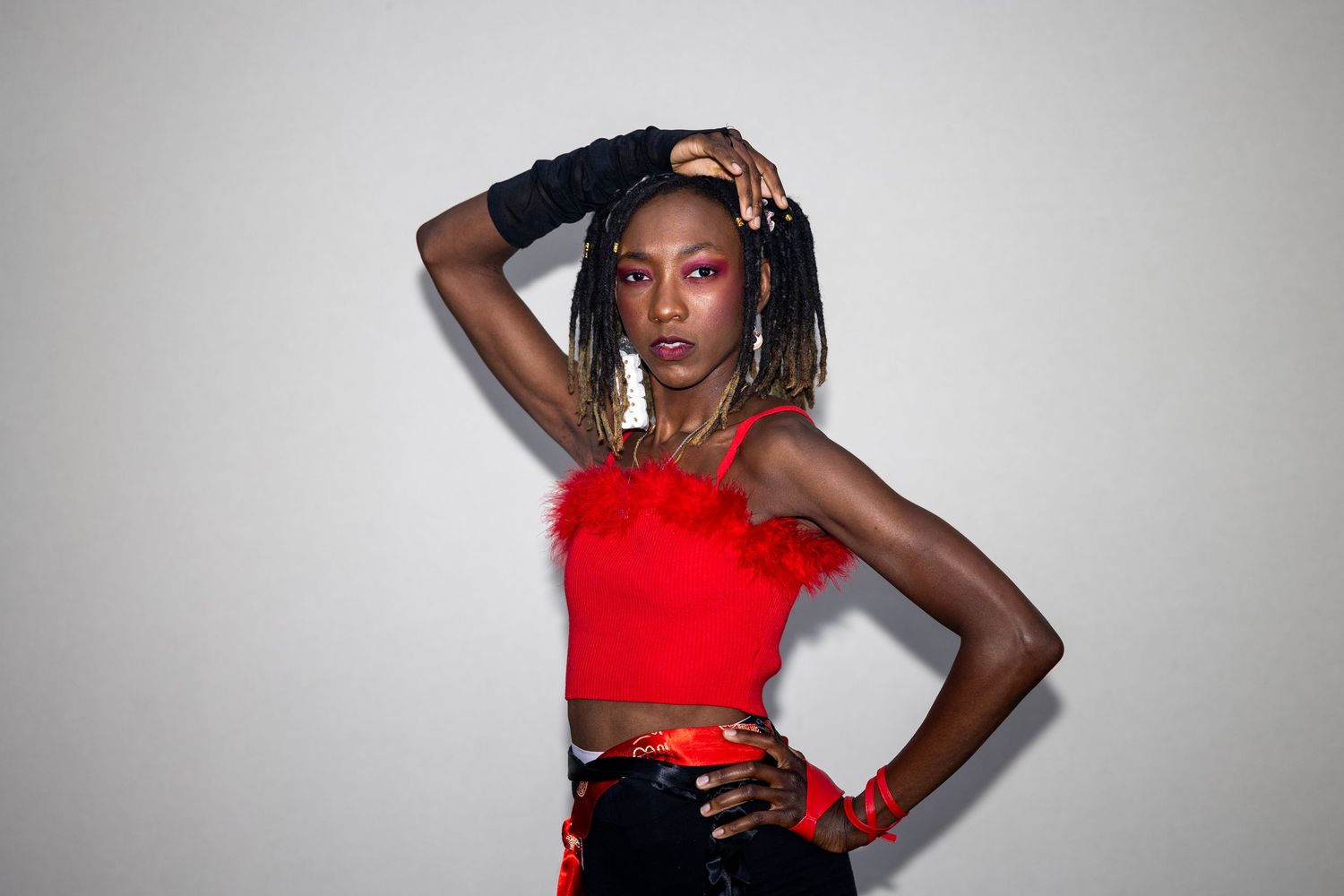 Young woman in red feathered crop top with braids poses confidently against white wall.