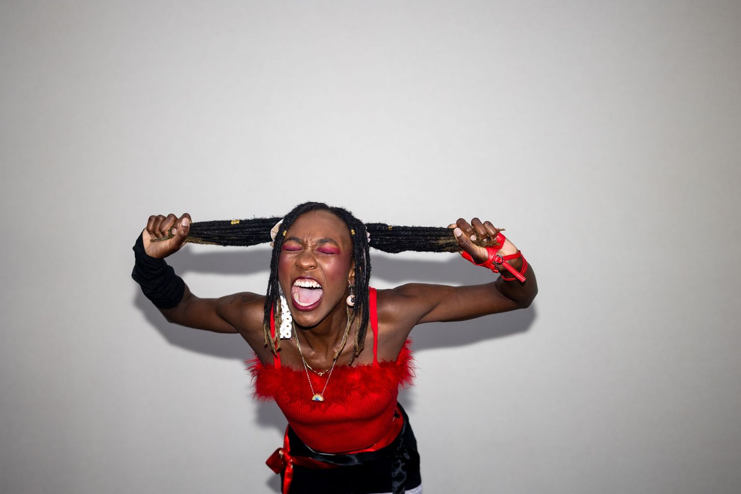 Energetic performer with braids in a red feathered crop top and black gloves pulling her hair while screaming.