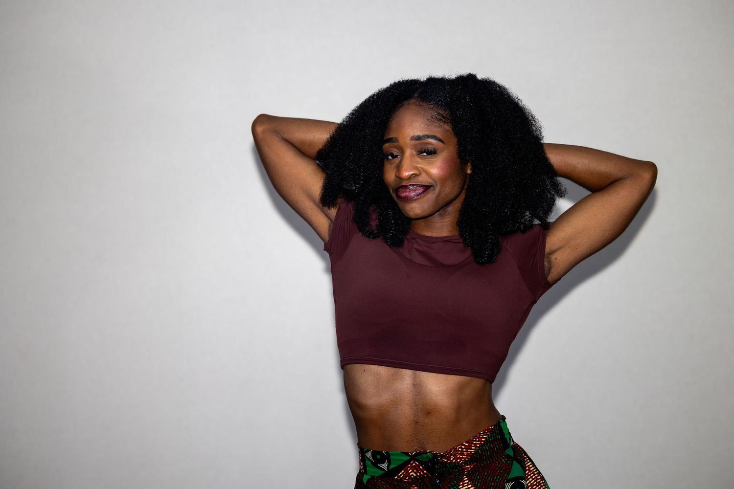 Smiling young Black woman with natural curly hair wearing a maroon crop top and printed skirt posing indoors.
