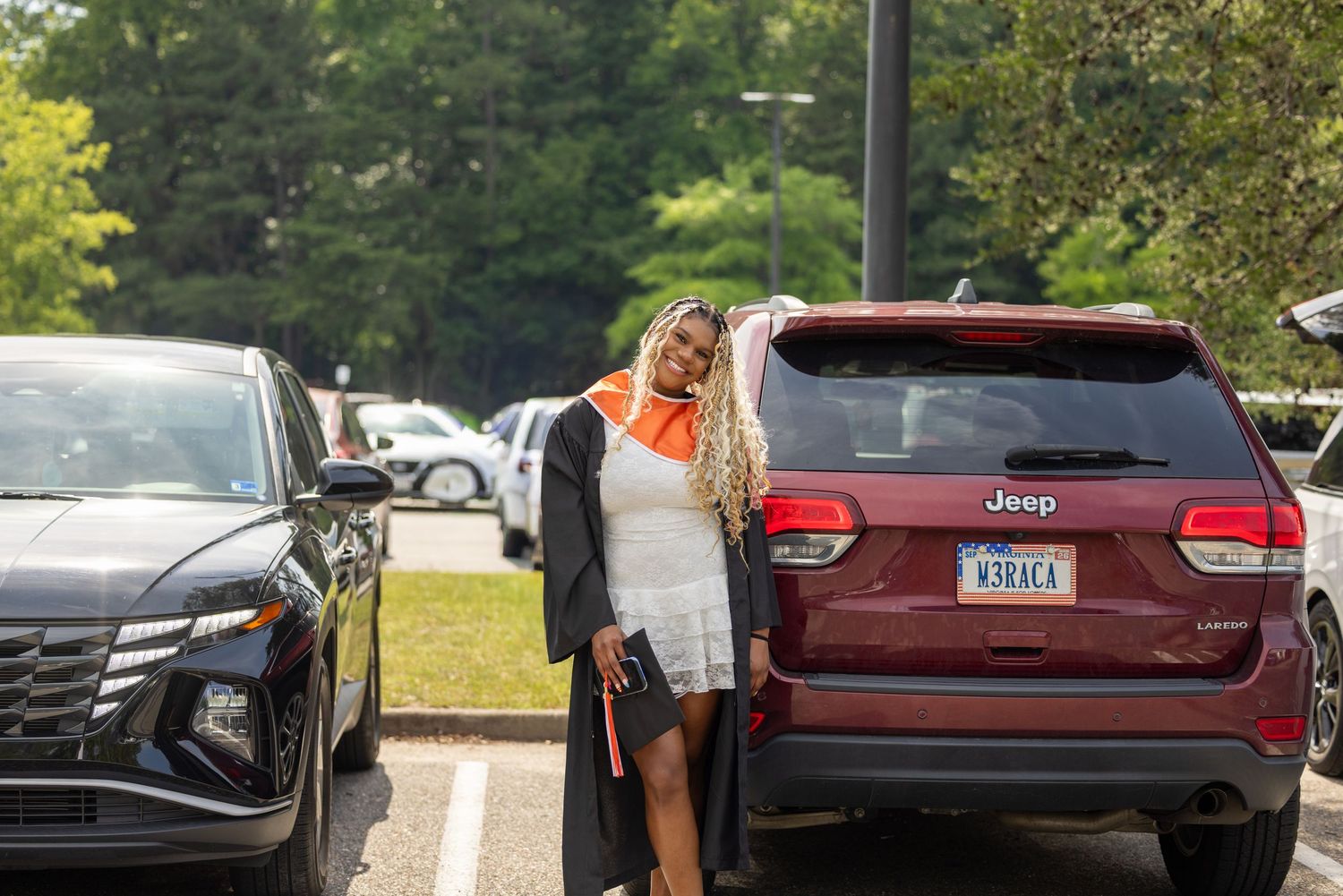 Graduate in cap and gown posing proudly beside a Jeep SUV in a parking lot on graduation day.