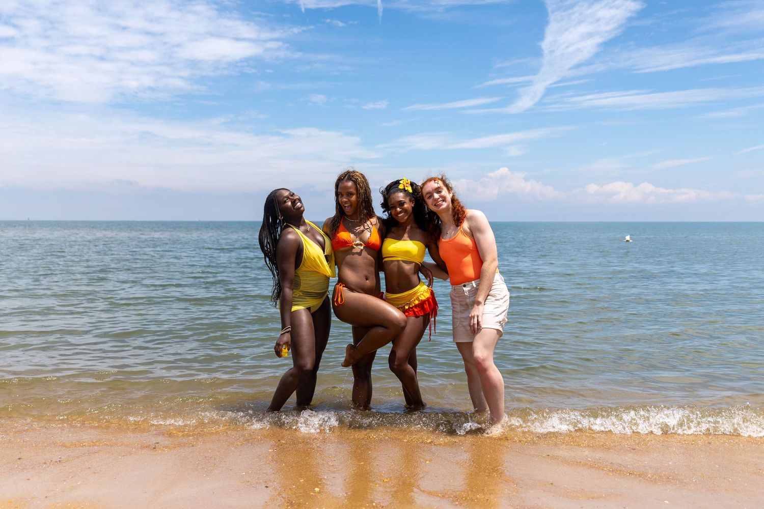 Four women in colorful swimsuits posing together at the beach on a sunny day.