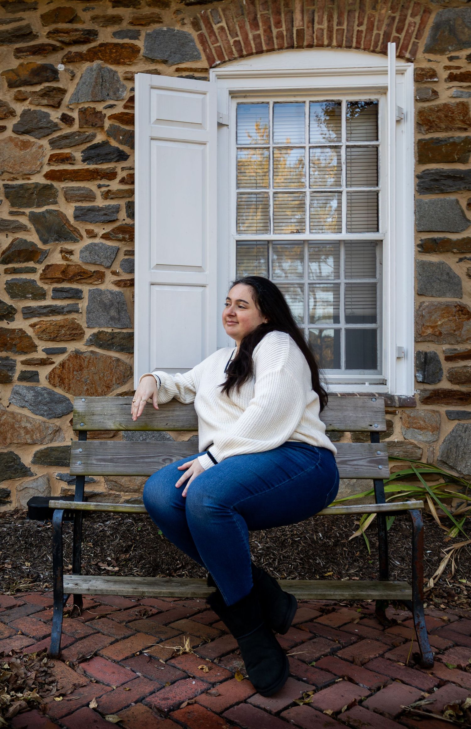 A person in white sweater and jeans poses playfully on a bench against a stone building wall.