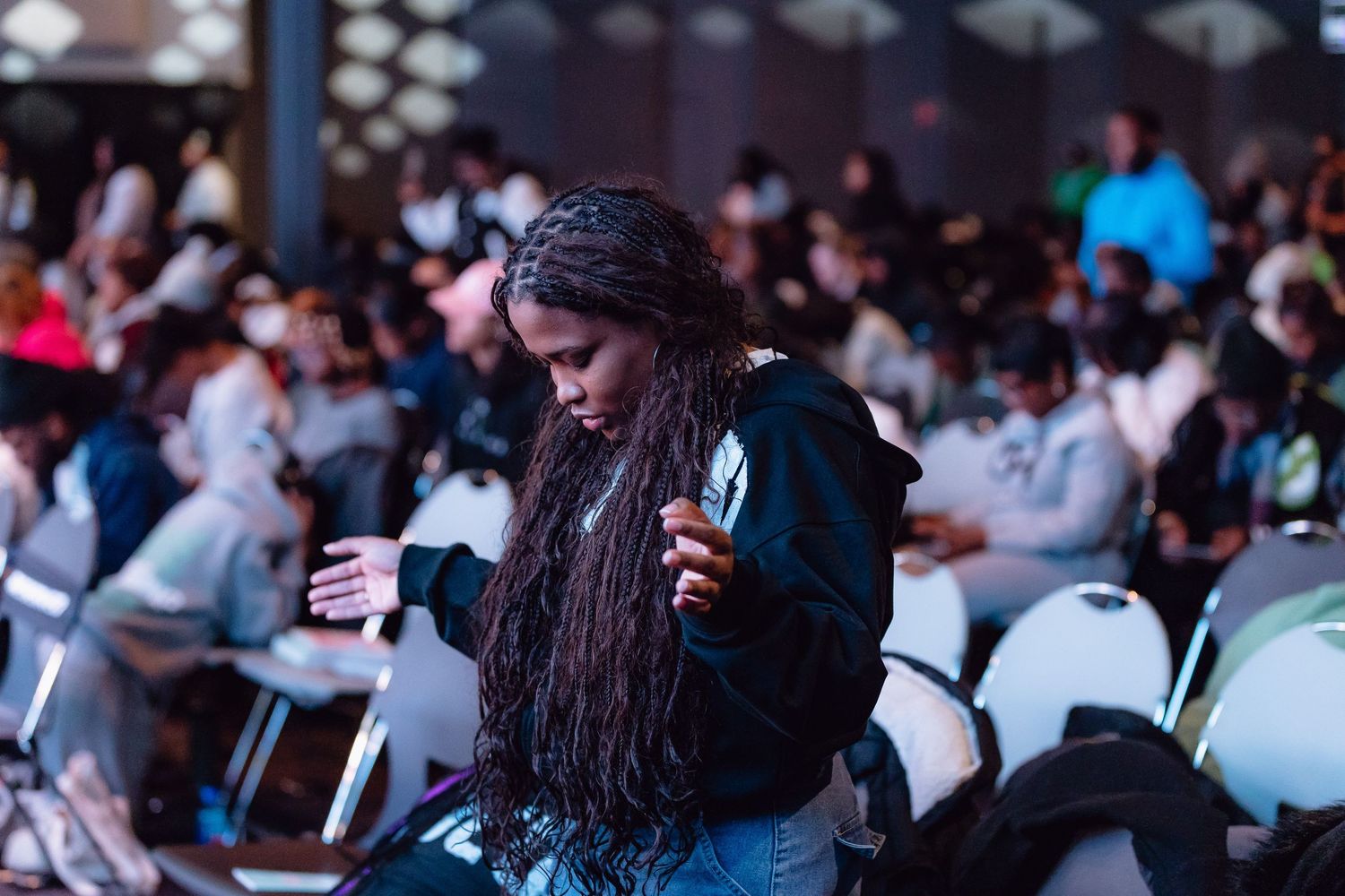 A performer with long dreadlocks gestures emotionally while speaking into a microphone at a crowded indoor event.