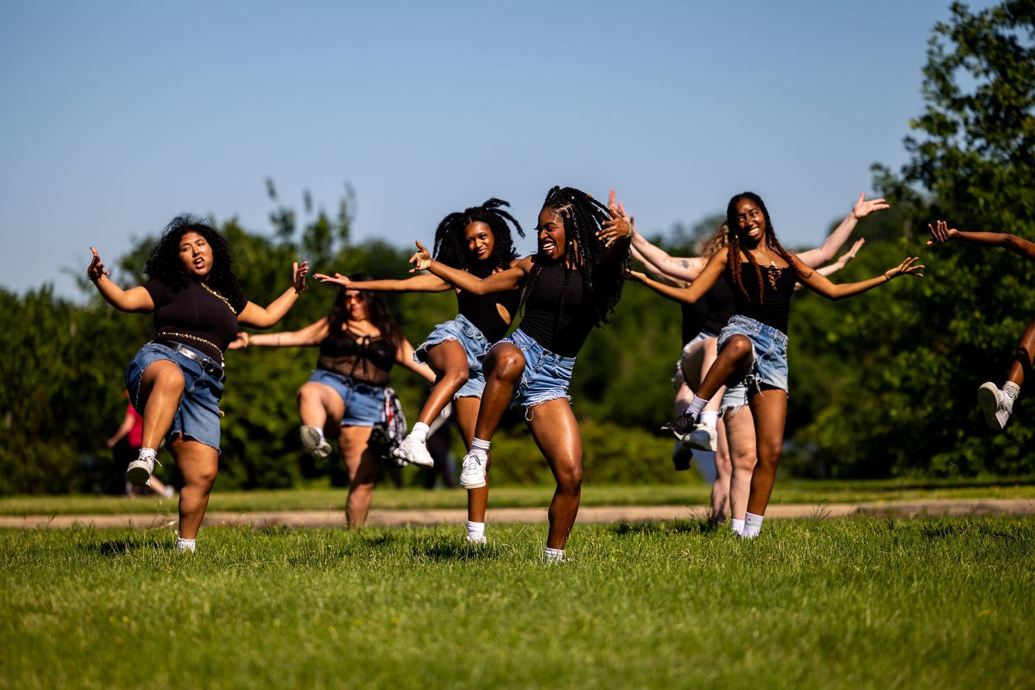 Five young women joyfully jumping and dancing together on a green lawn under a clear blue sky.