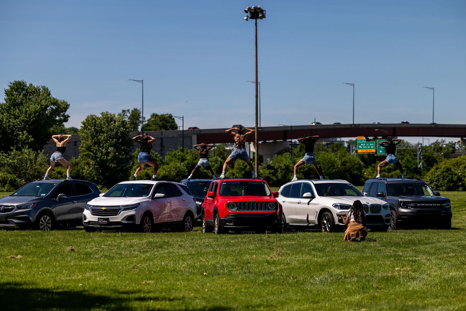 Multiple SUVs and Jeeps parked on green grass near a highway interchange on a sunny day.