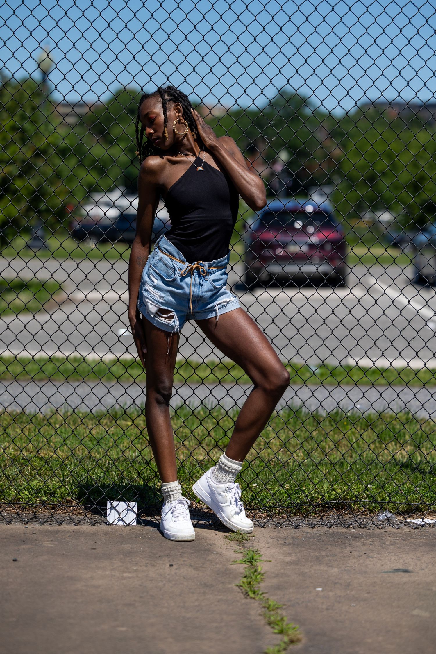 Young Black woman in denim shorts and white sneakers posing against a chain-link fence outdoors.