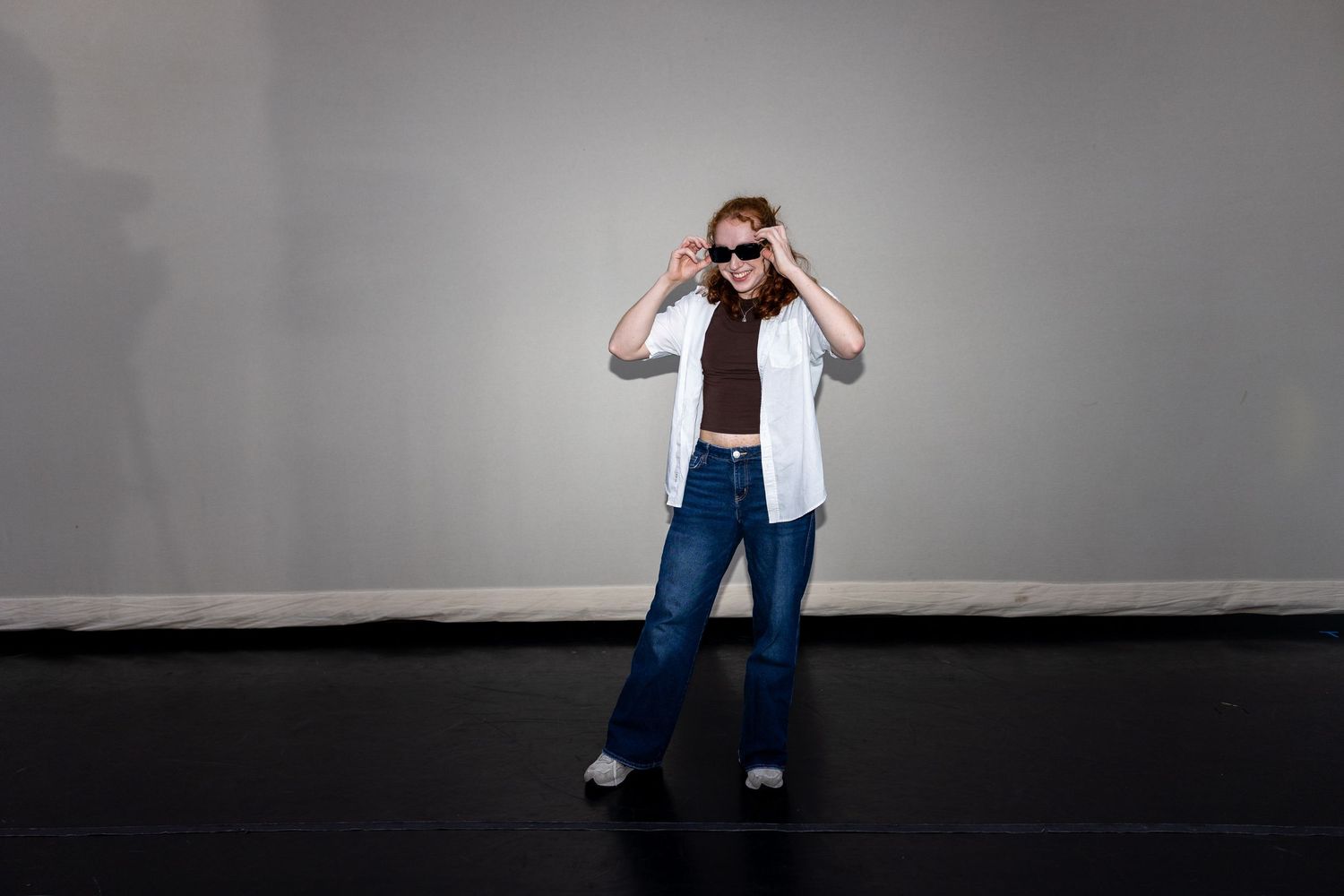 Redhead woman in sunglasses posing in white shirt and flared jeans on dark stage.
