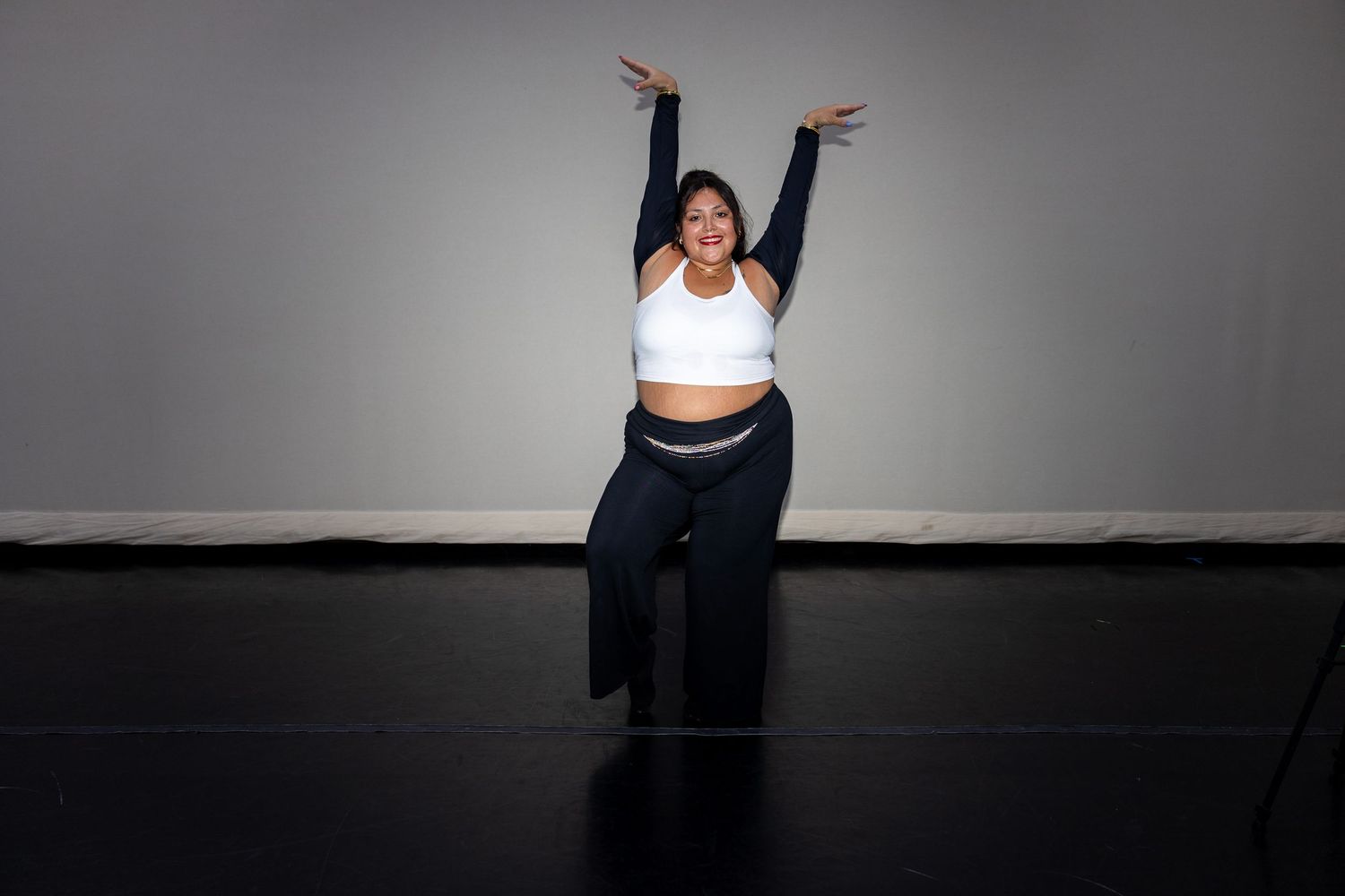 A joyful dancer raises her arms in a white sports bra and navy pants on a black studio floor.
