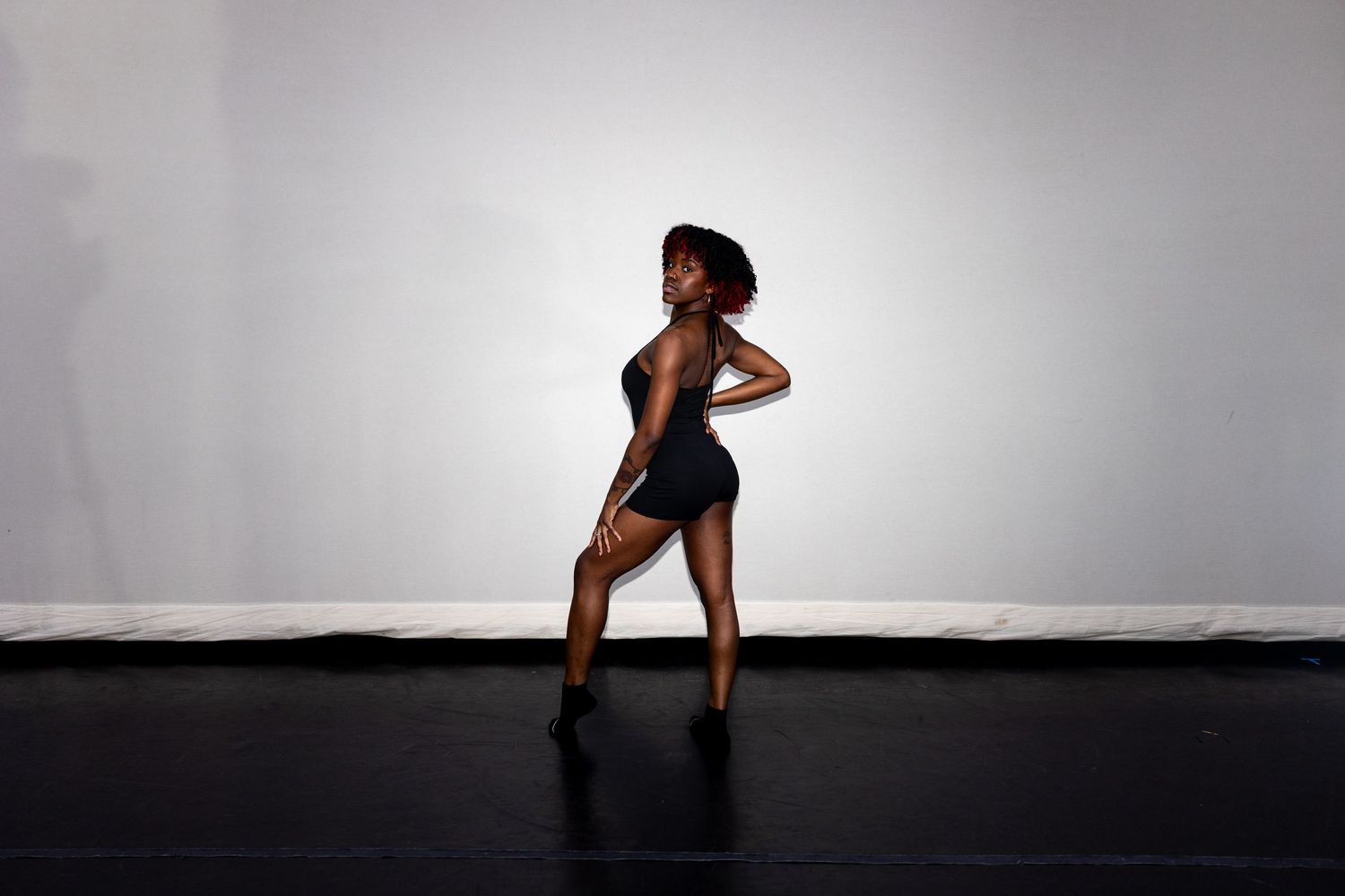 A dancer in a black bodysuit and heels poses confidently on a dark studio floor.