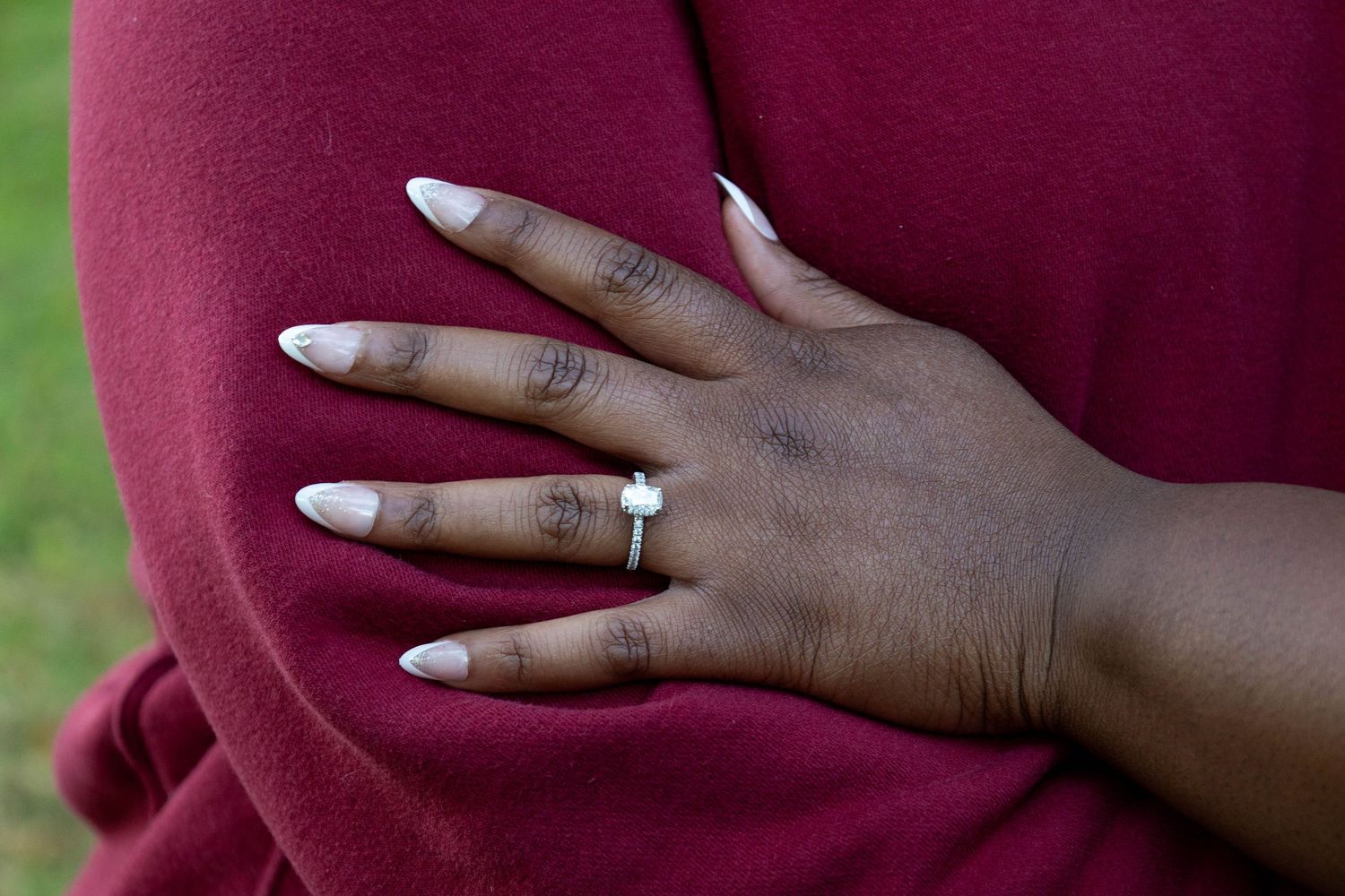 Close-up of a woman's hand displaying a diamond engagement ring with long white nails on a red sweater.