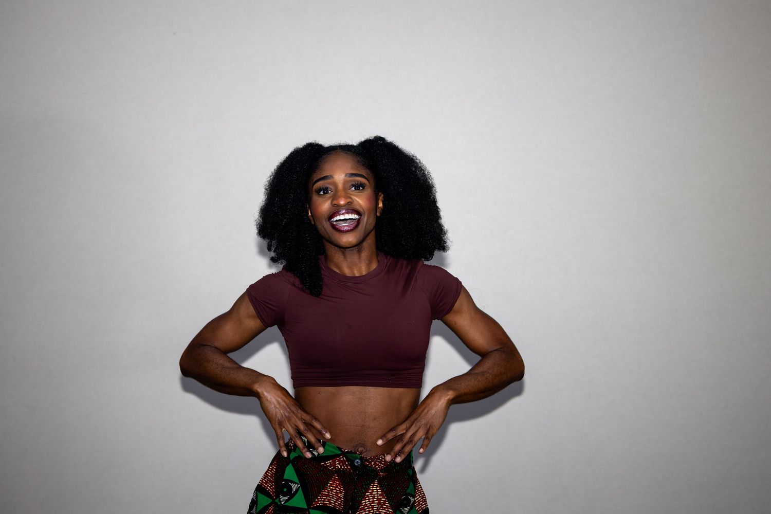 Young woman with natural curly hair wearing a maroon crop top and colorful skirt posing with hands on hips.