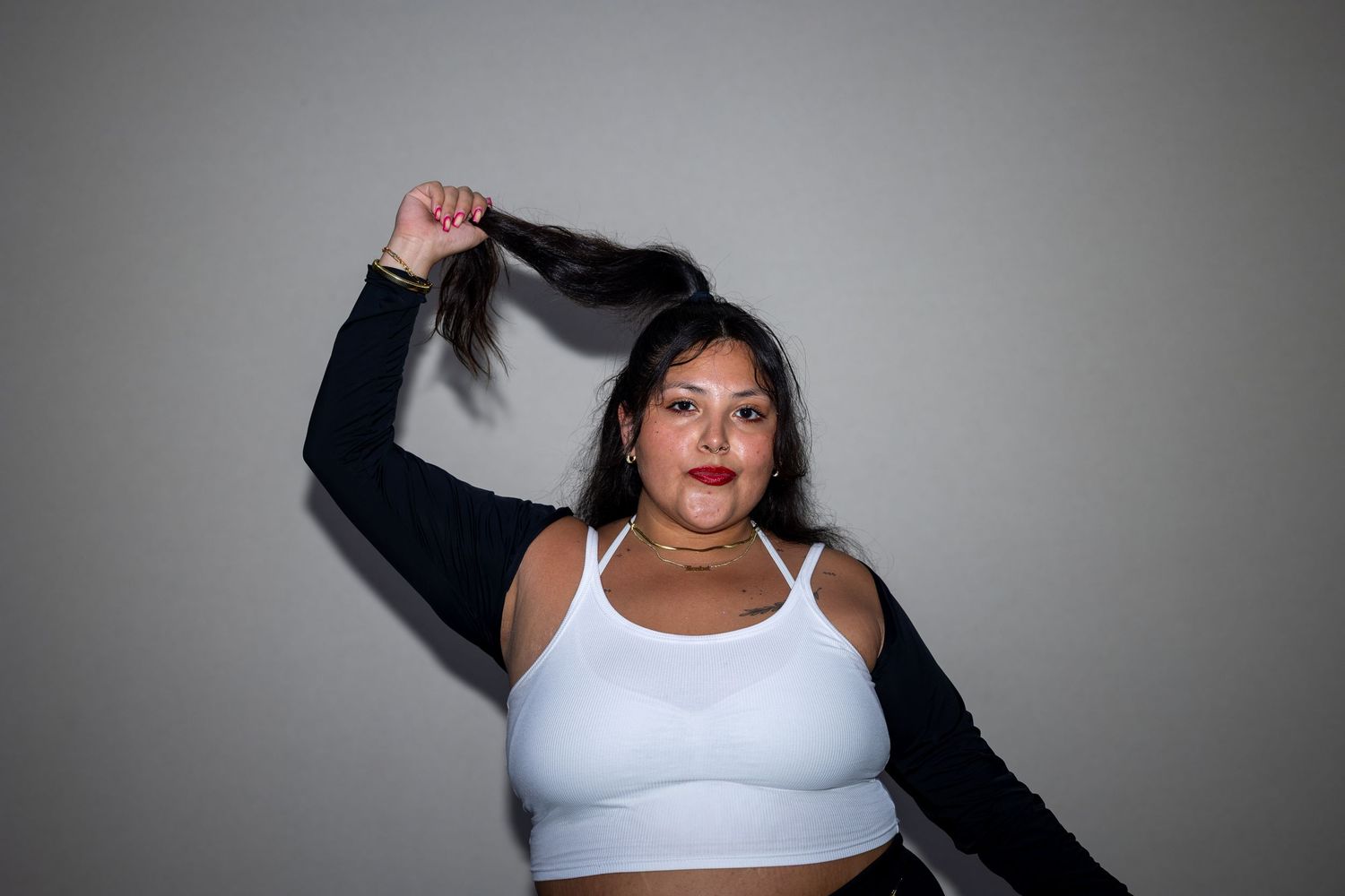 Woman in white crop top and black jacket holds her dark hair up against a gray background.