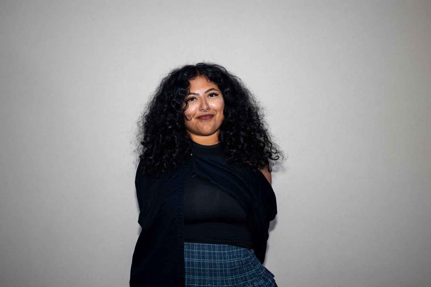 Young woman with curly dark hair wearing a black off-shoulder top and plaid skirt smiles warmly.