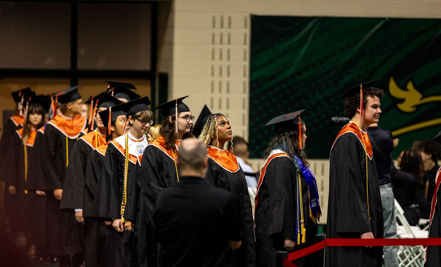 Graduates in black caps and gowns with orange sashes stand in line during a graduation ceremony indoors.