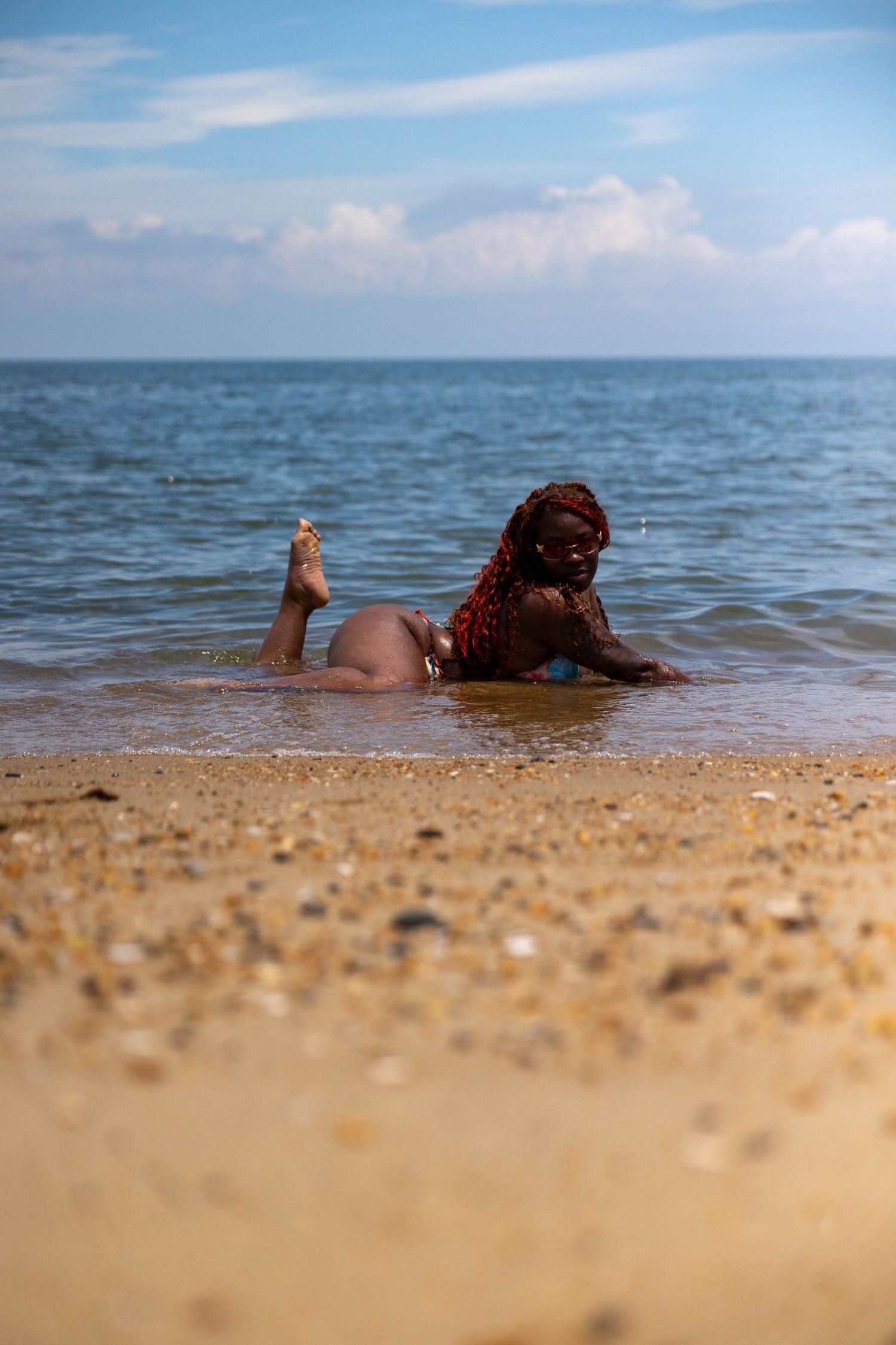 A Black woman with red braids in a bikini lies on a sandy beach shore with ocean waves behind her.