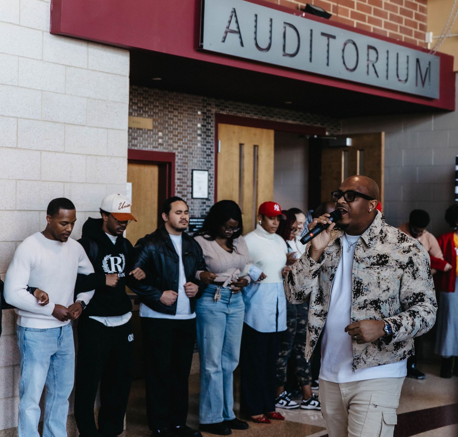People waiting in line outside an auditorium entrance along a brick wall.