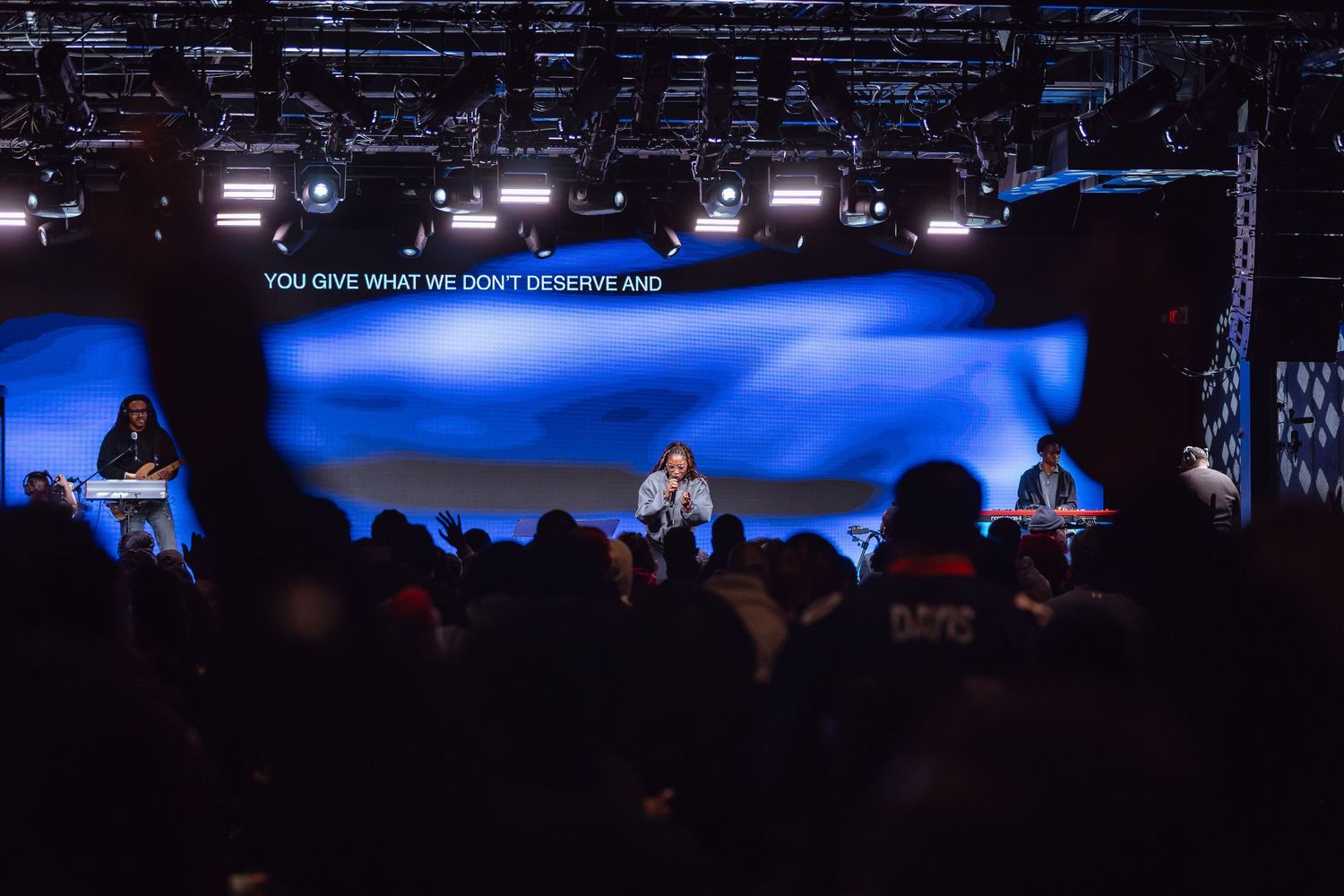 Concert stage with blue lighting and silhouetted crowd visible in a dark venue during a live performance.