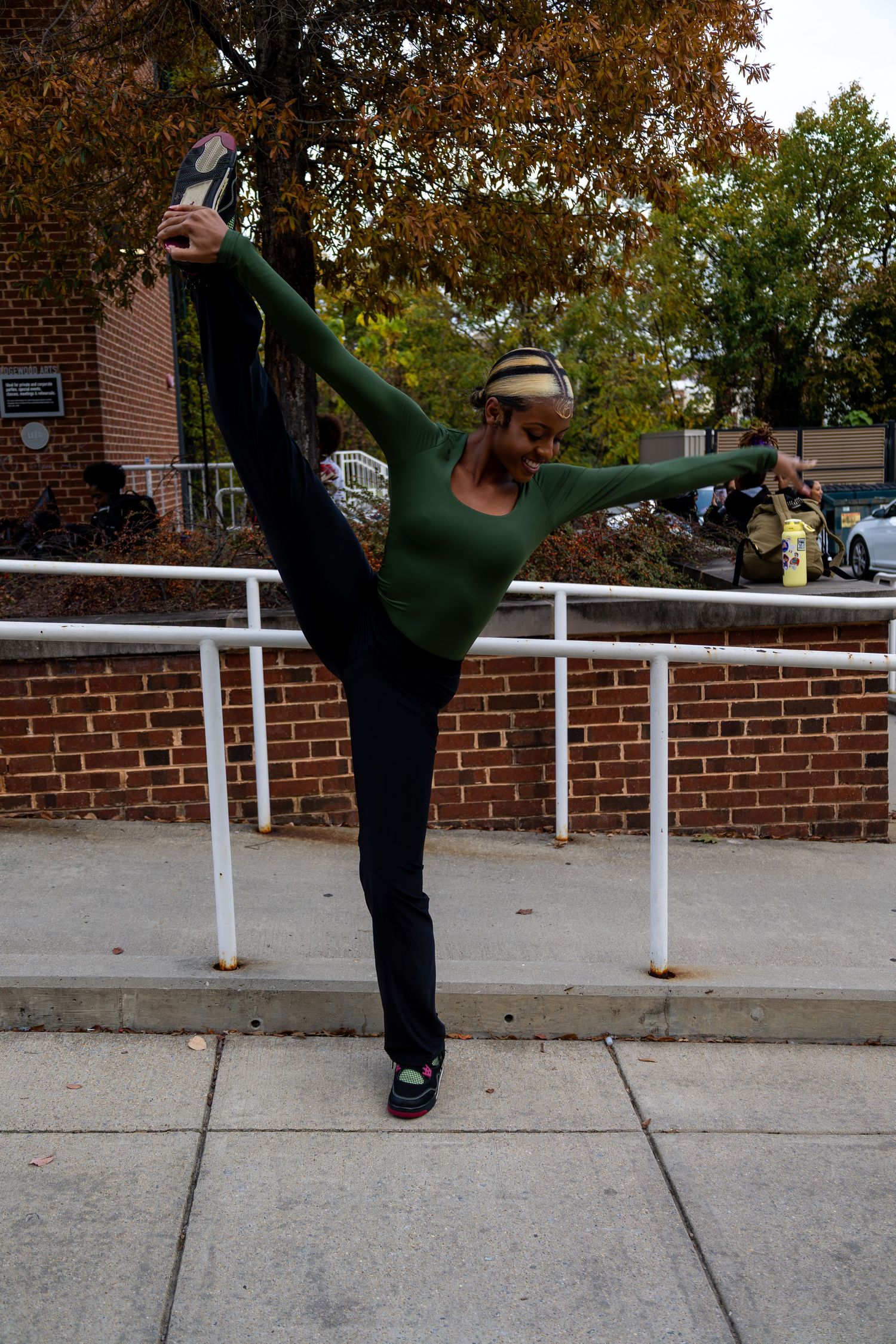 Person in green top executes high kick pose by white railing on concrete path.