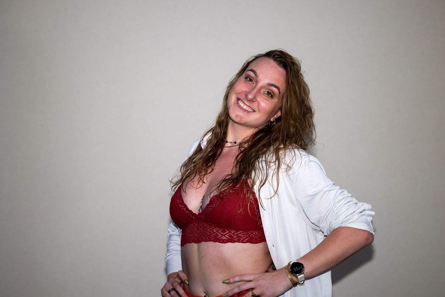 Woman in red lace bralette and open white shirt smiling confidently against a light gray background.