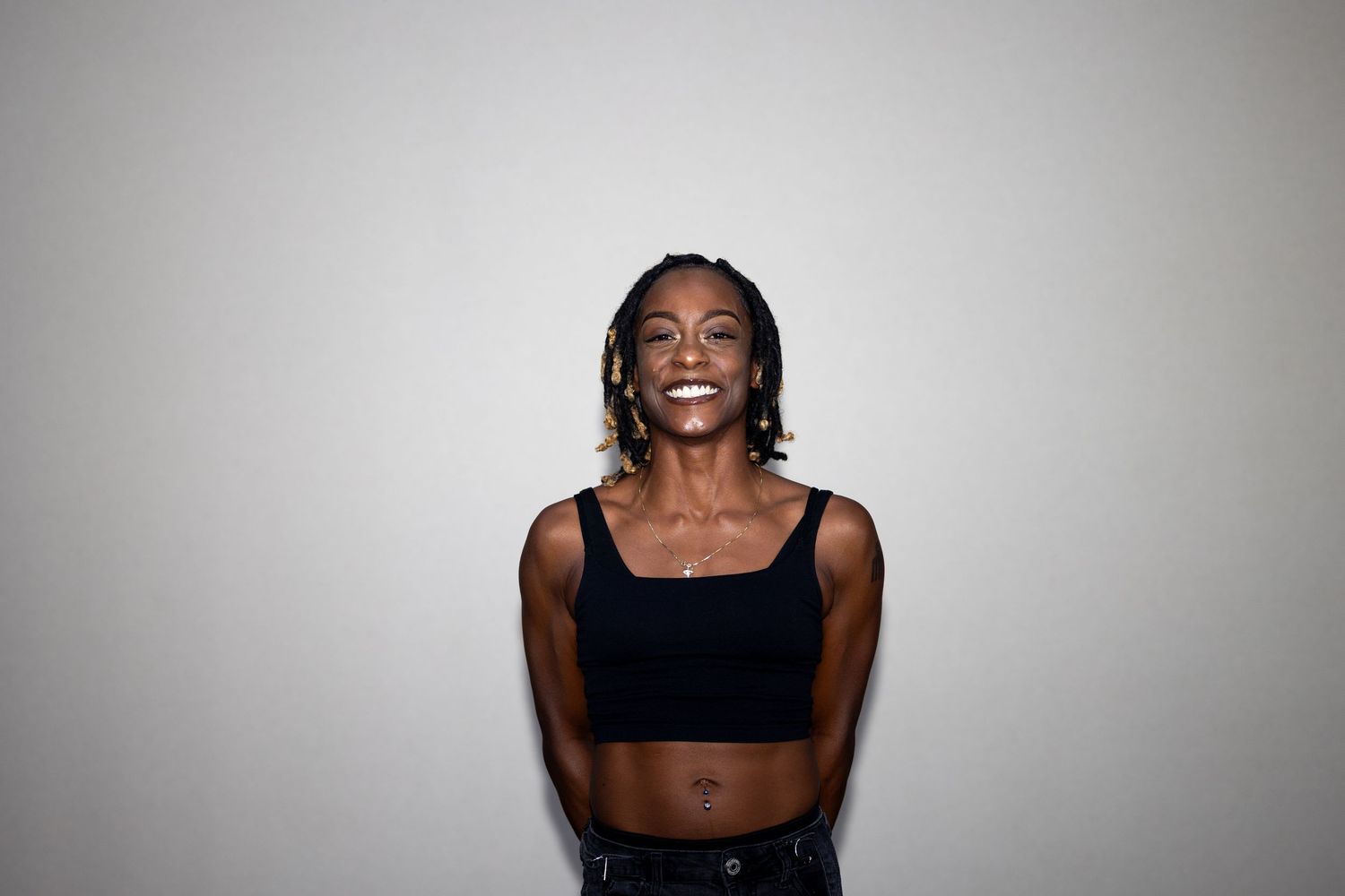 A slender Black woman with braids laughs wearing a black square-neck crop top against white wall.