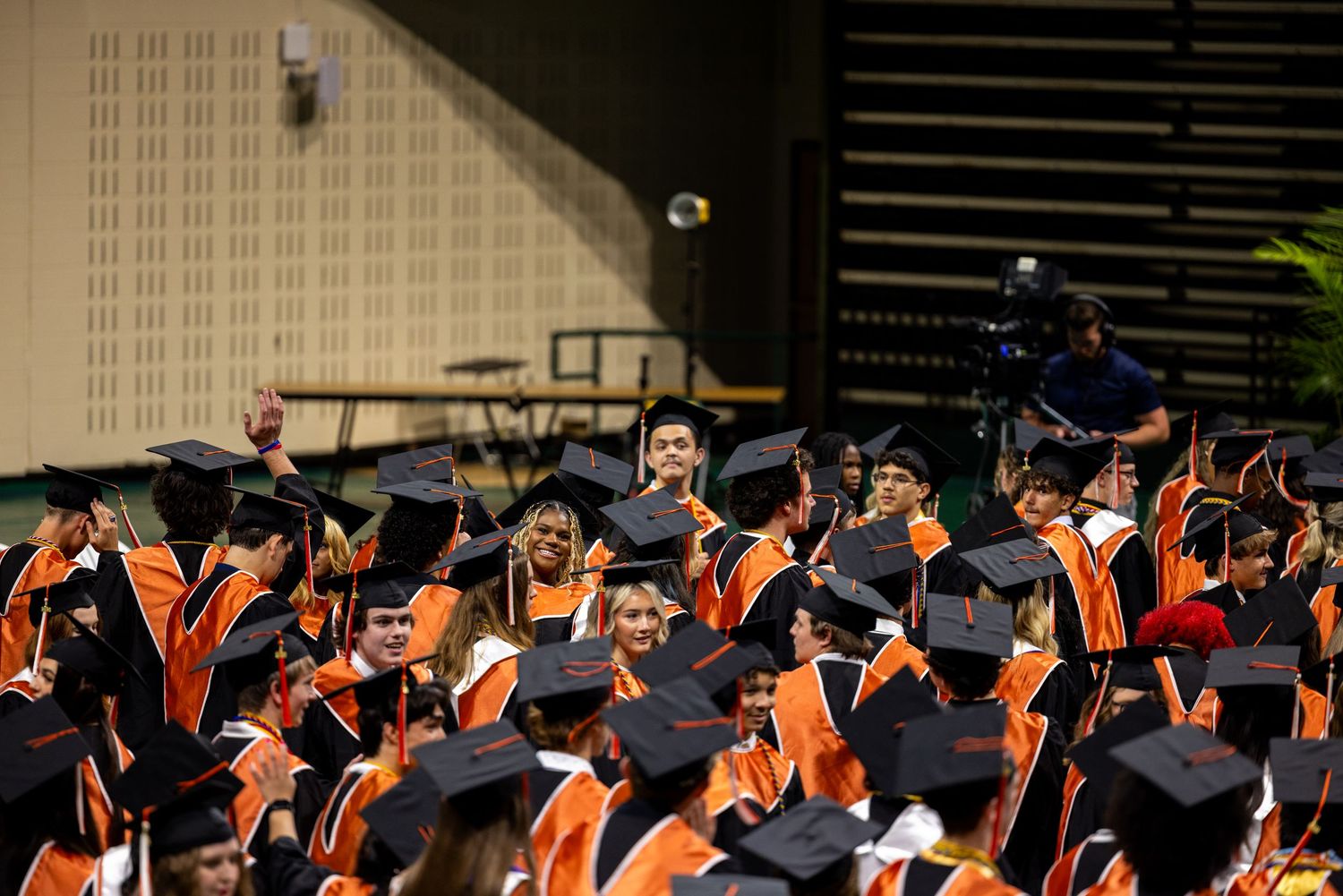 Graduates in black caps and orange robes celebrate at a university commencement ceremony indoors.