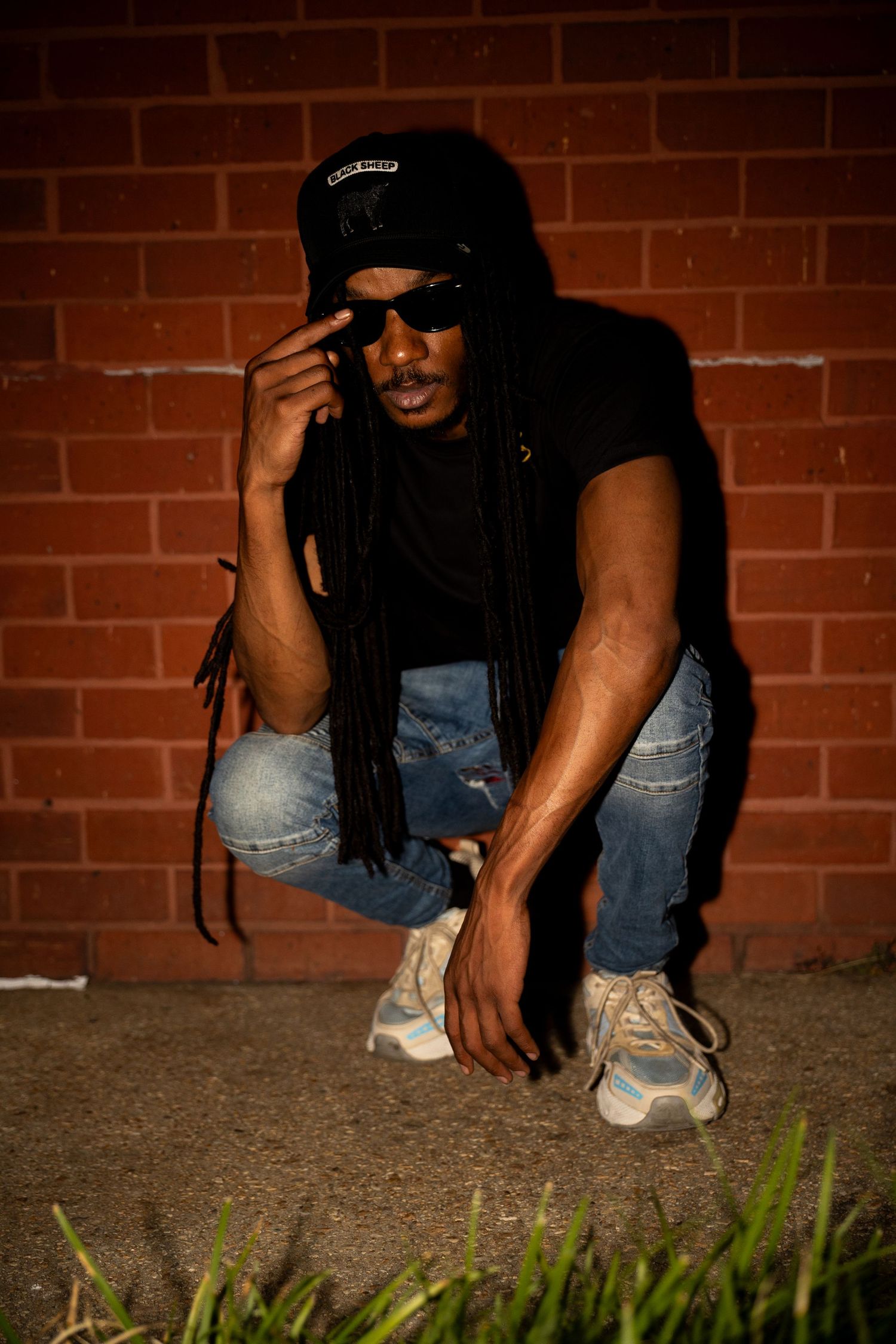Man with long dreadlocks crouching by brick wall, wearing black cap, sunglasses, and ripped jeans.