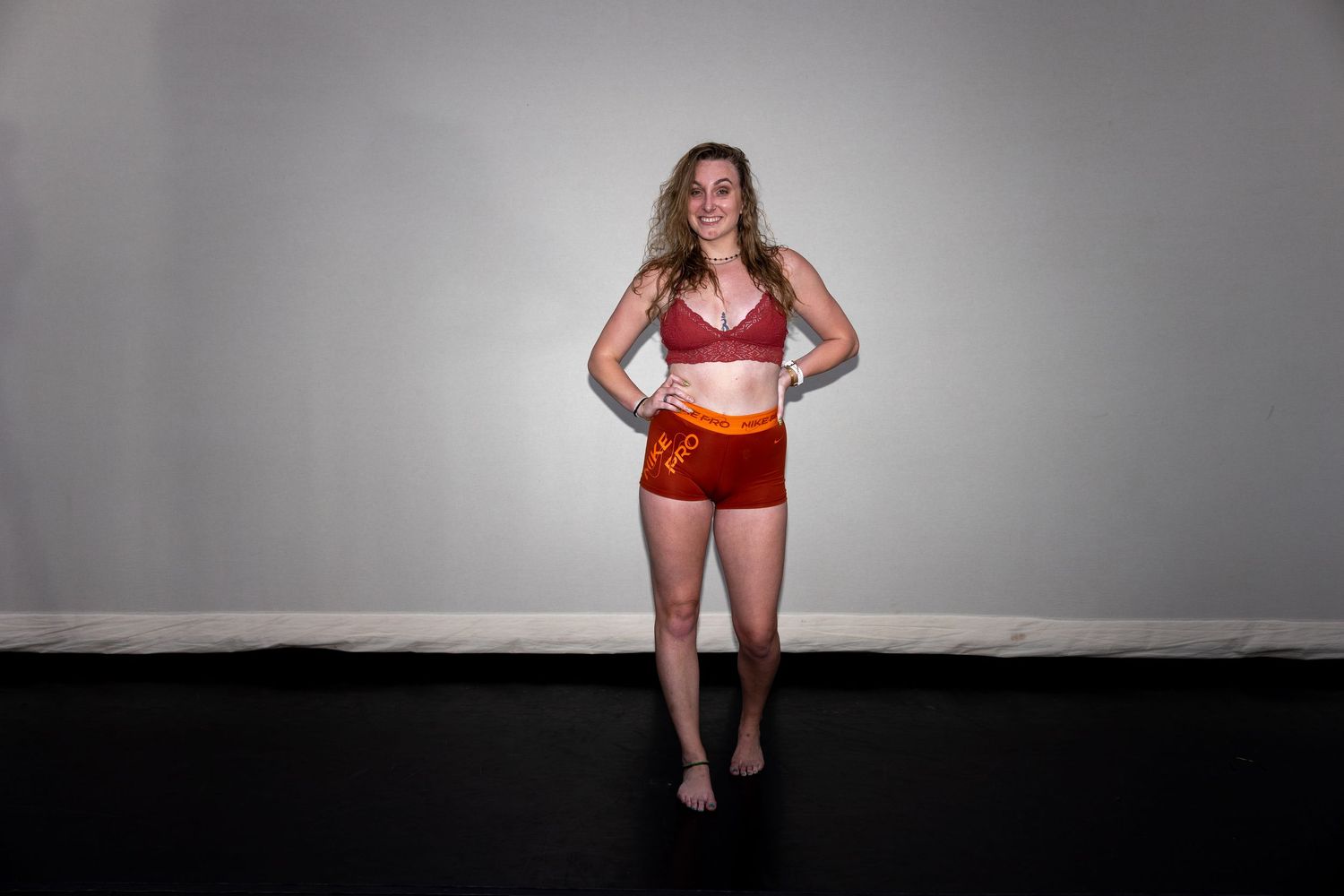 Woman posing in red crop top and orange shorts against a gray wall on a black floor.