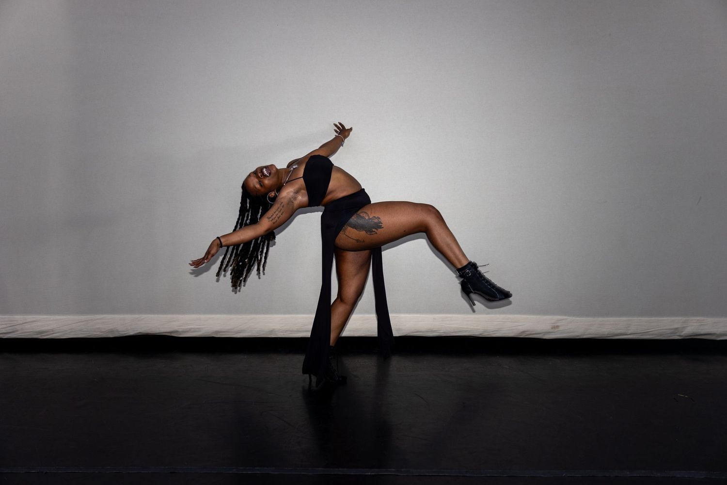 Black female dancer performs dramatic backbend in black outfit and heels on studio floor.