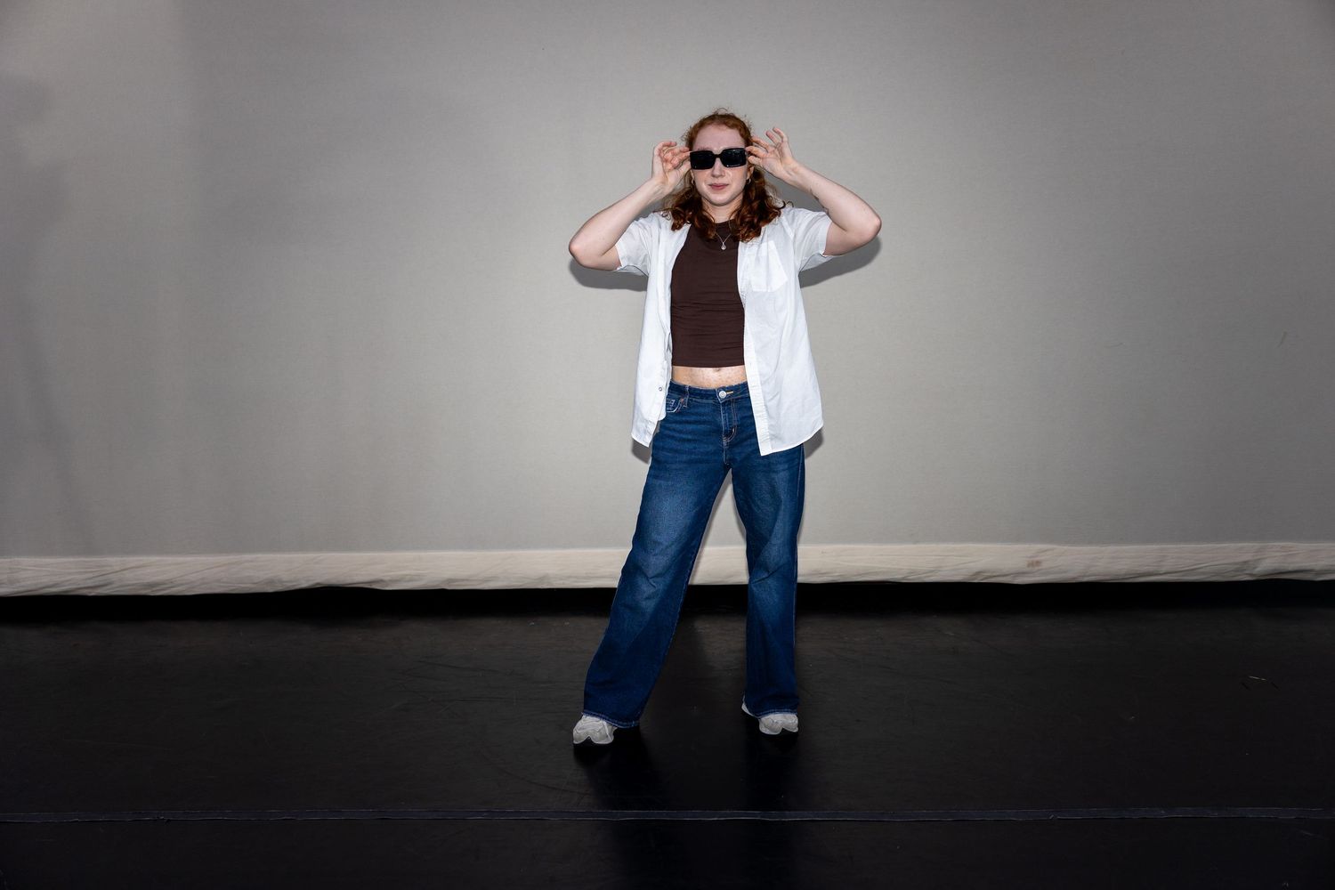 Woman in white shirt and jeans adjusting sunglasses while posing in dance studio.